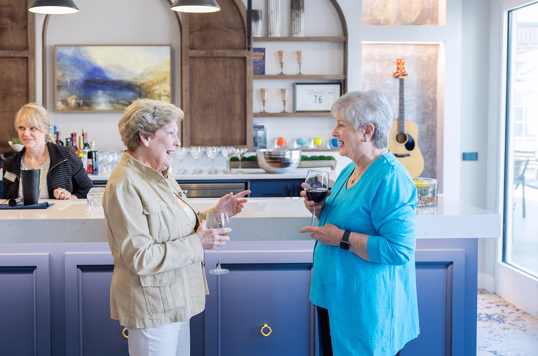 Two women stand by a counter, smiling and holding wine glasses while talking.