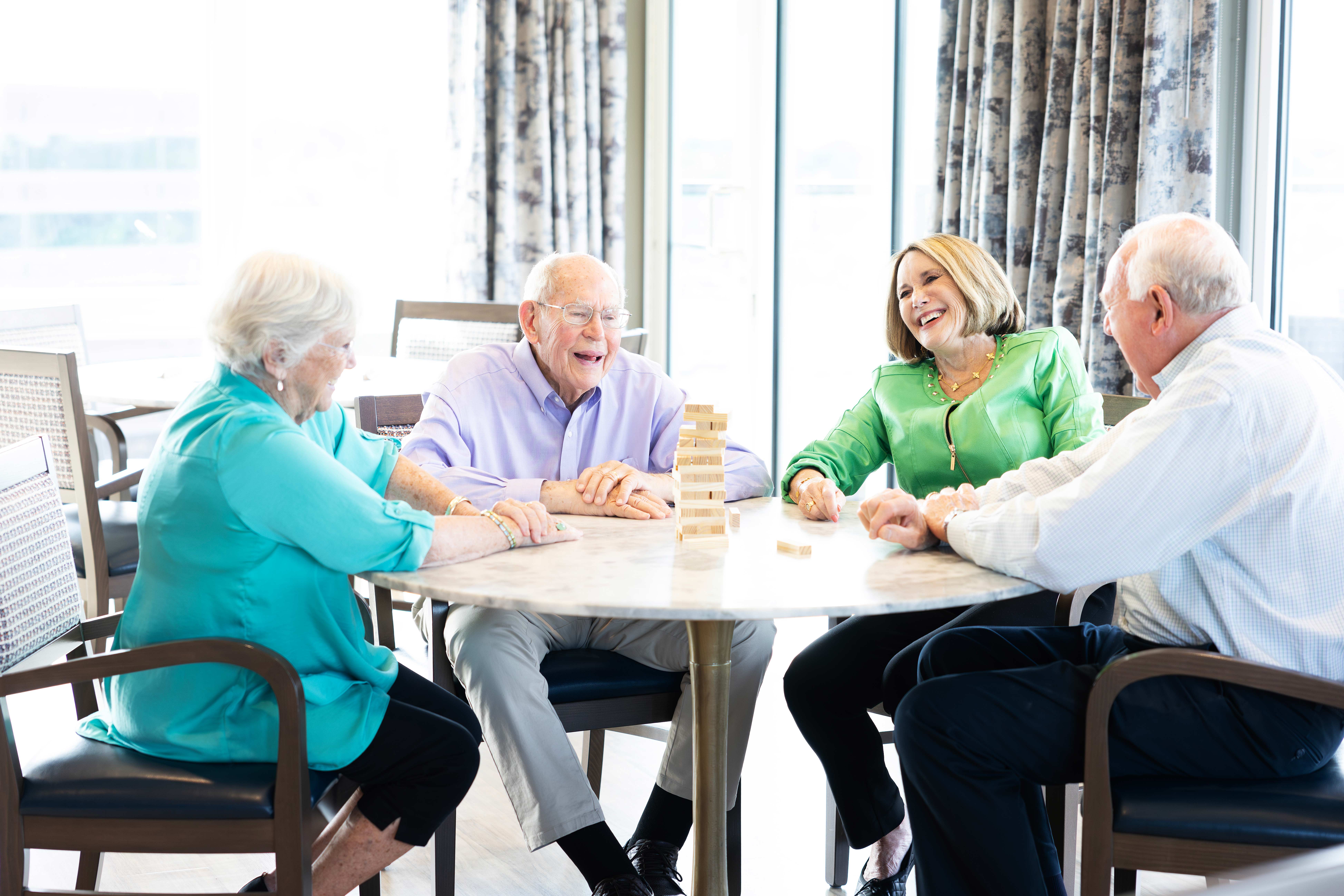 Four older adults are sitting around a table, playing Jenga and laughing together.