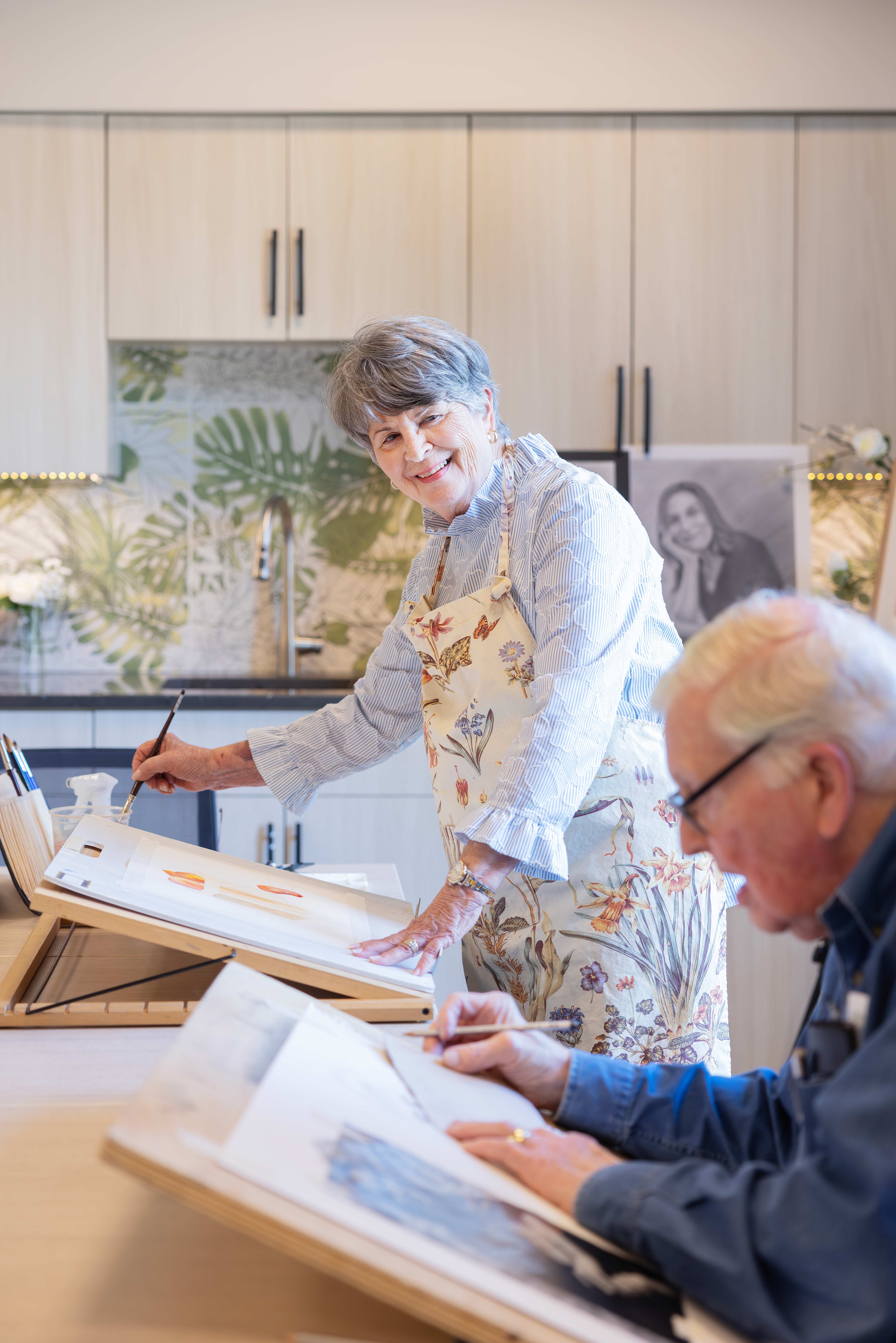 Two elderly individuals are painting at a table, smiling and focused on their artwork.