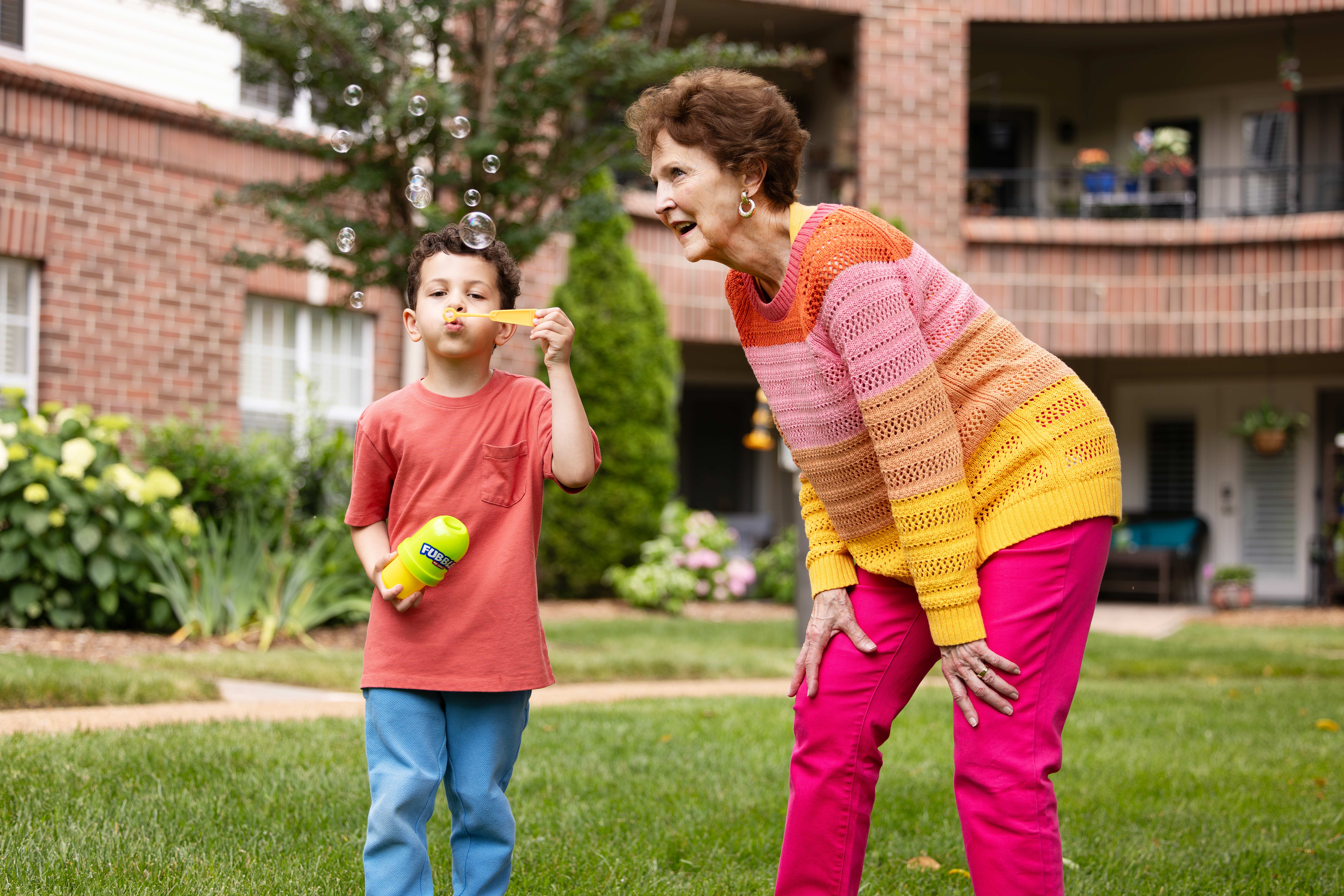 A child blows bubbles while an older woman watches and smiles in a grassy area.