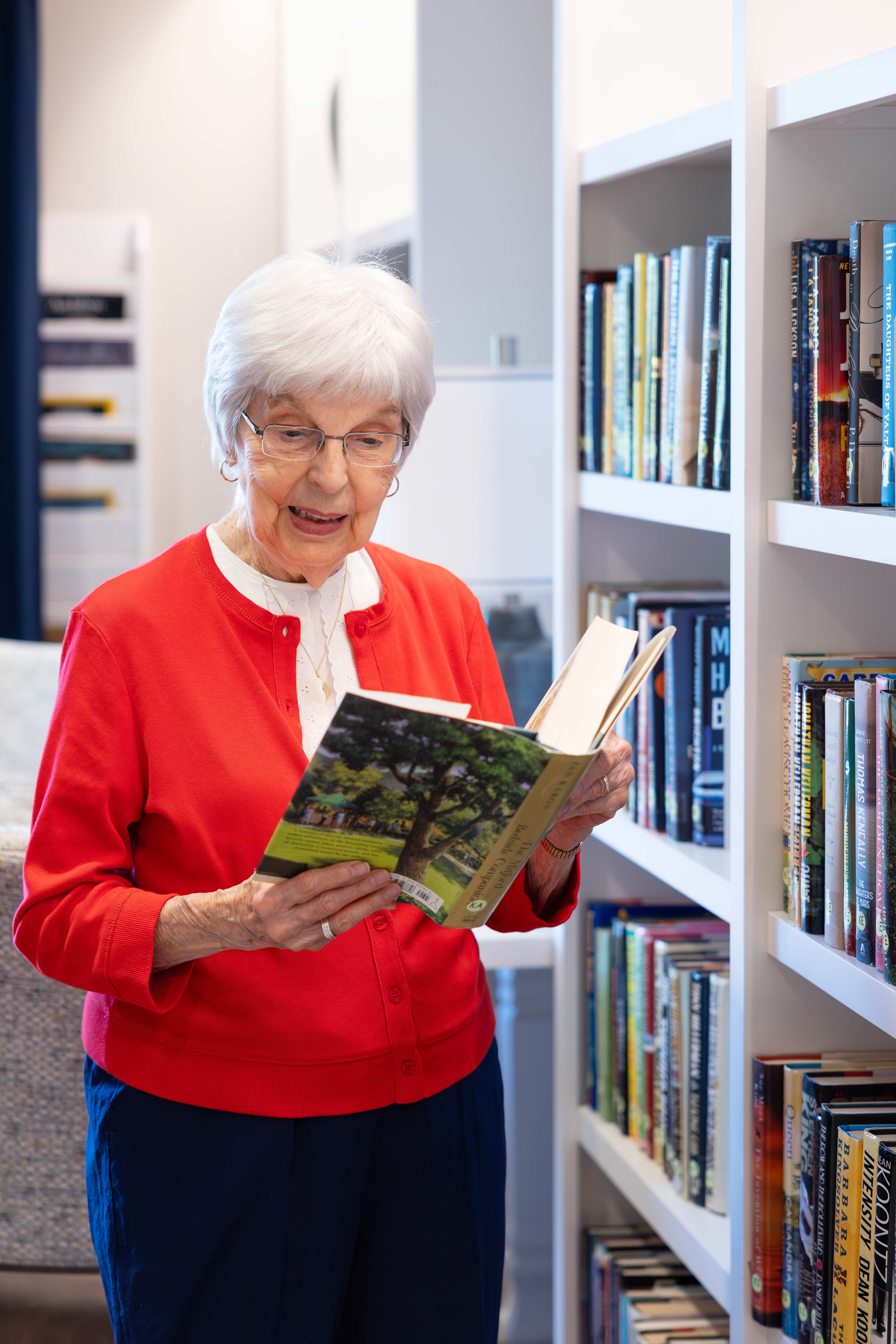 An elderly woman in a red sweater reads a book while standing in a library.
