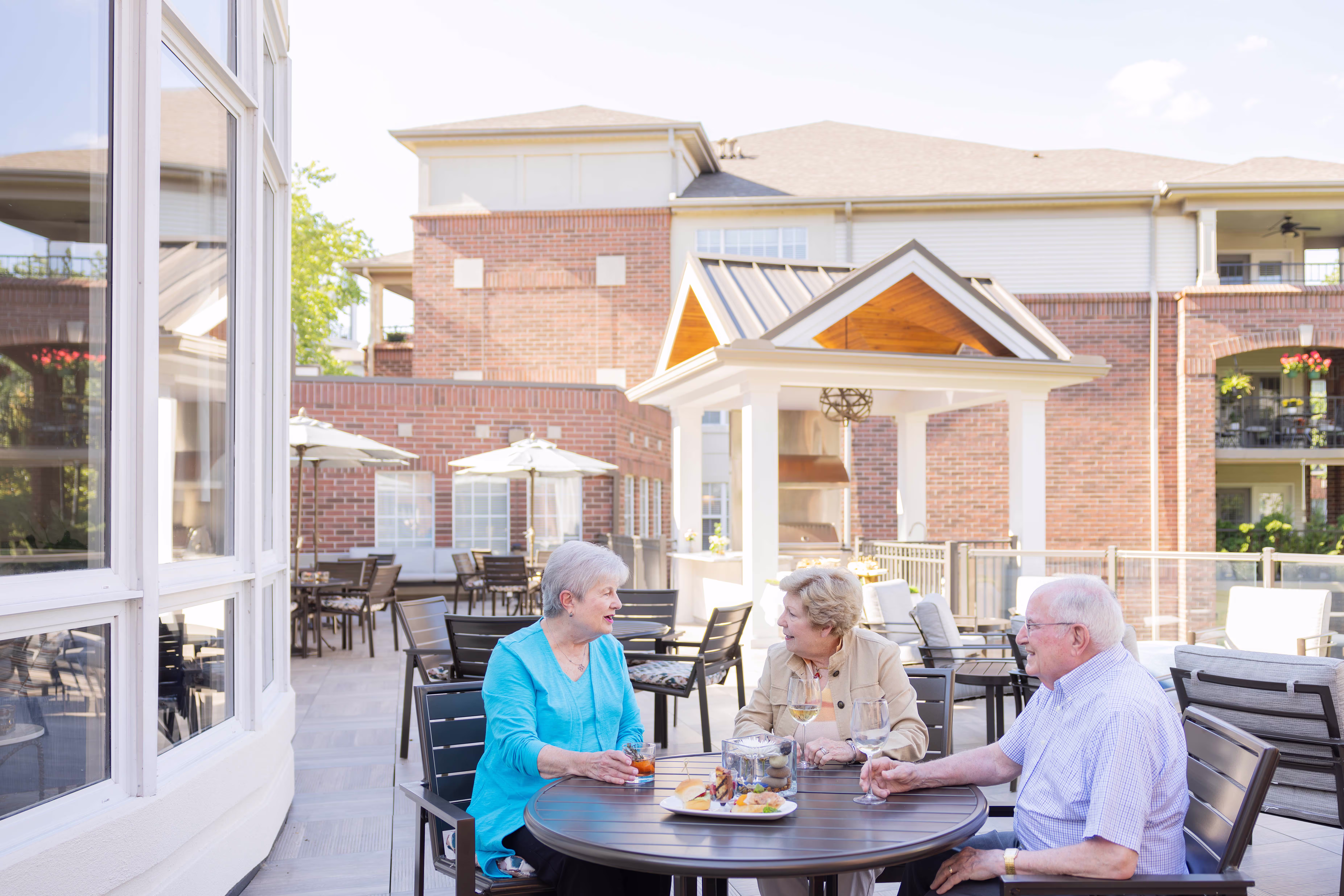 Three older adults sit at a round table outside, enjoying drinks and chatting together.