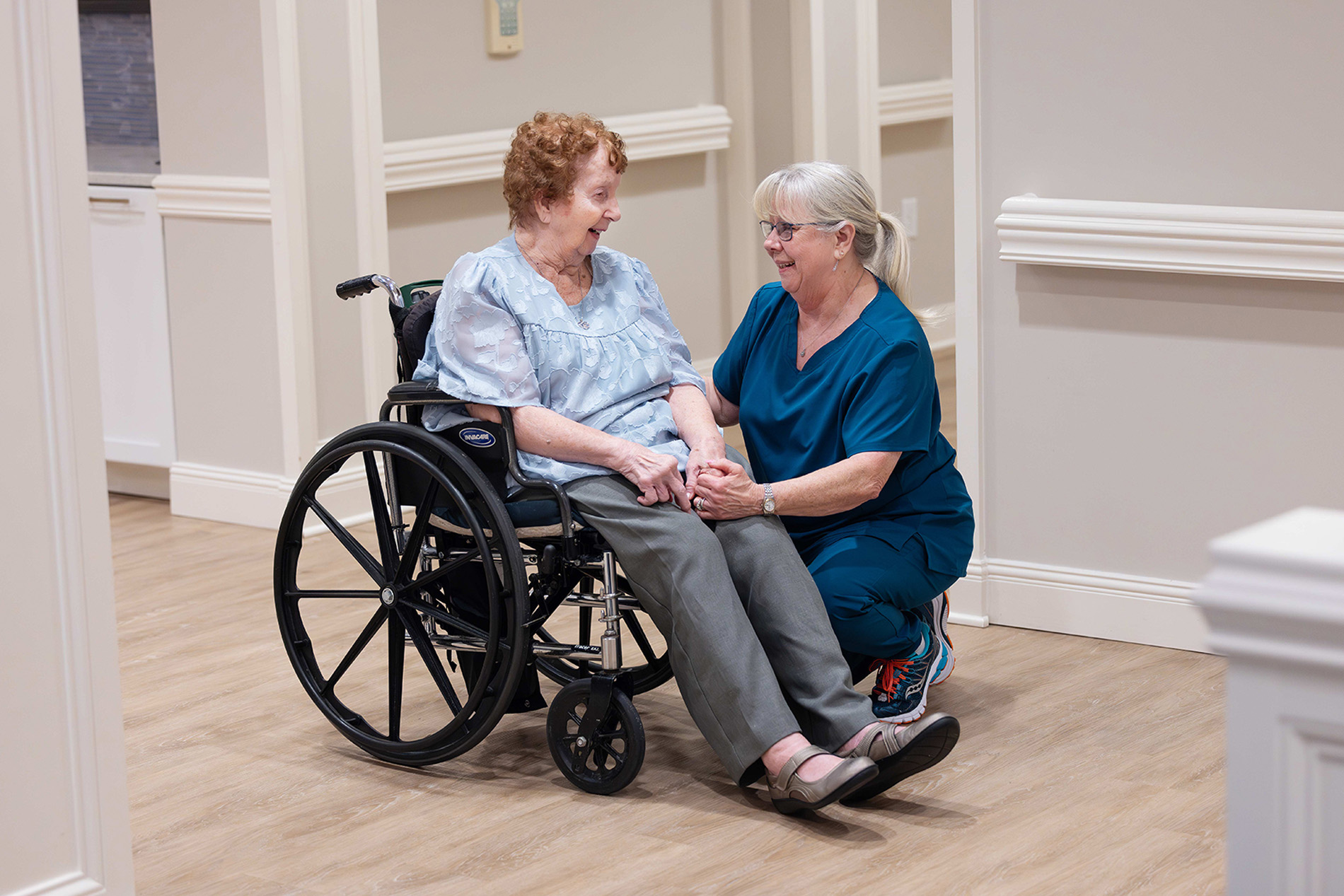 A nurse kneels and holds hands with an elderly woman sitting in a wheelchair, both smiling.