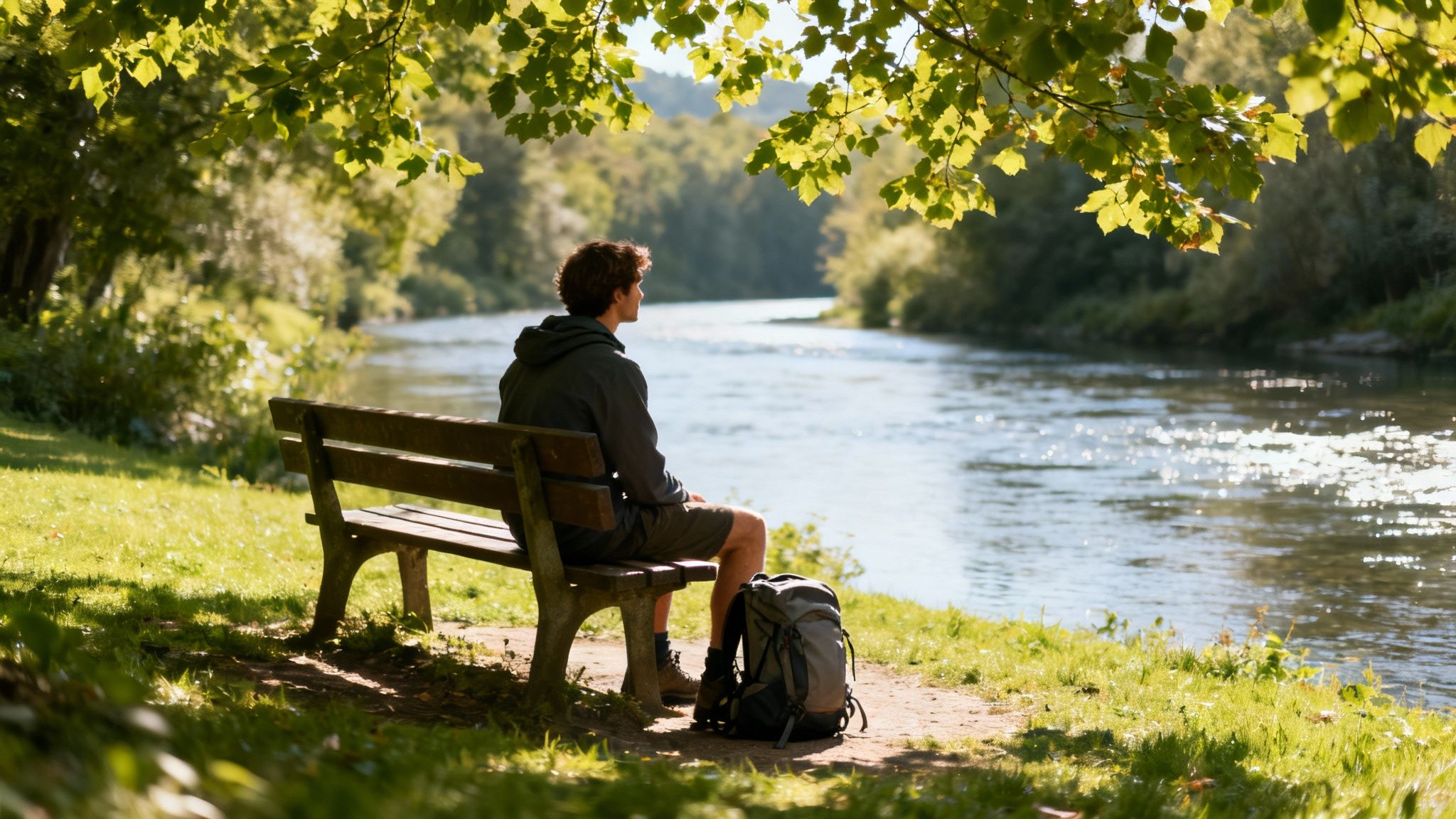 A man sits on a bench by a sunny river, contemplating nature with his backpack nearby.