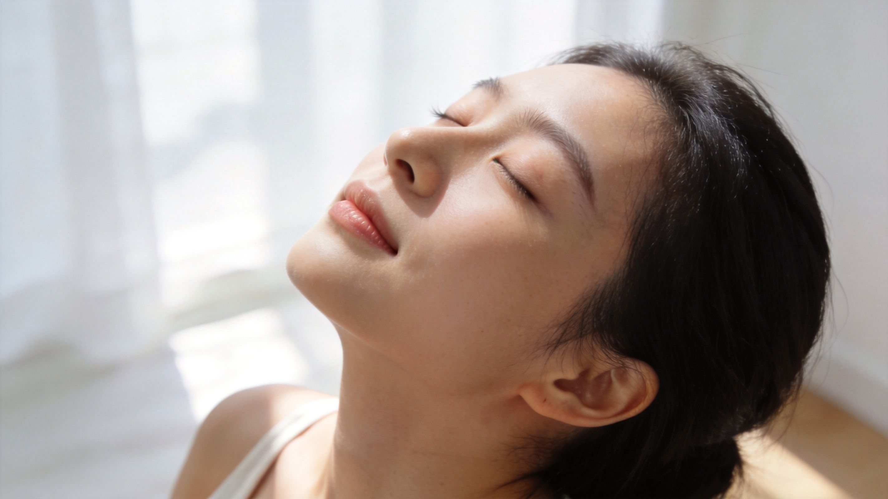 A young woman with glowing, clear skin basking in natural sunlight with her eyes closed peacefully.