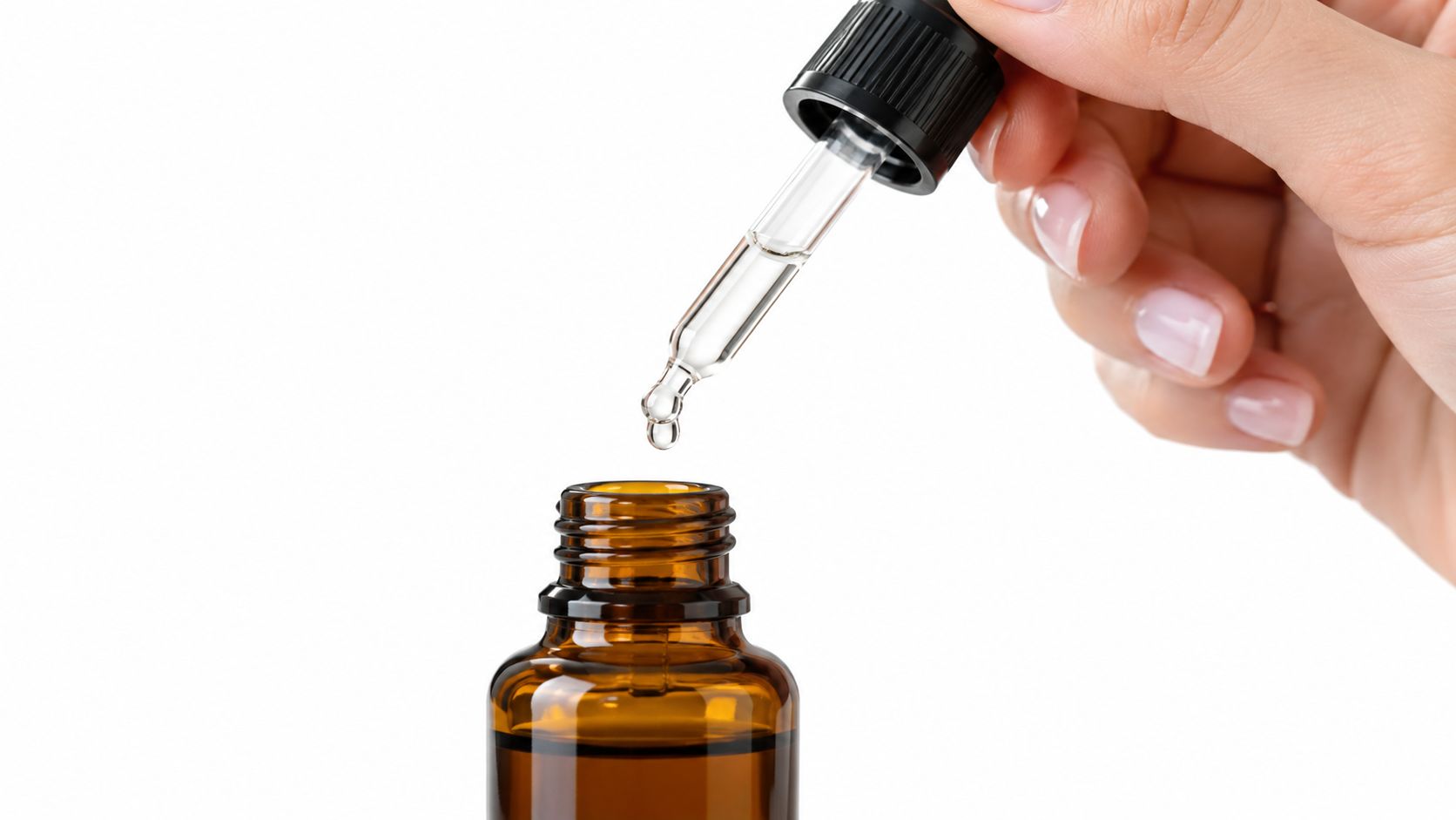 A hand holding a glass dropper dispensing a clear liquid into a small amber bottle isolated white.