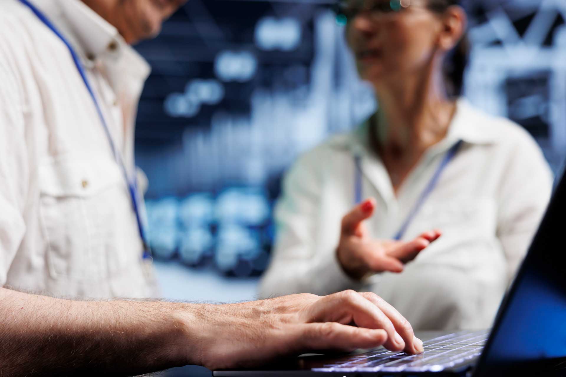 A person types on a laptop while another gestures during a conversation nearby.