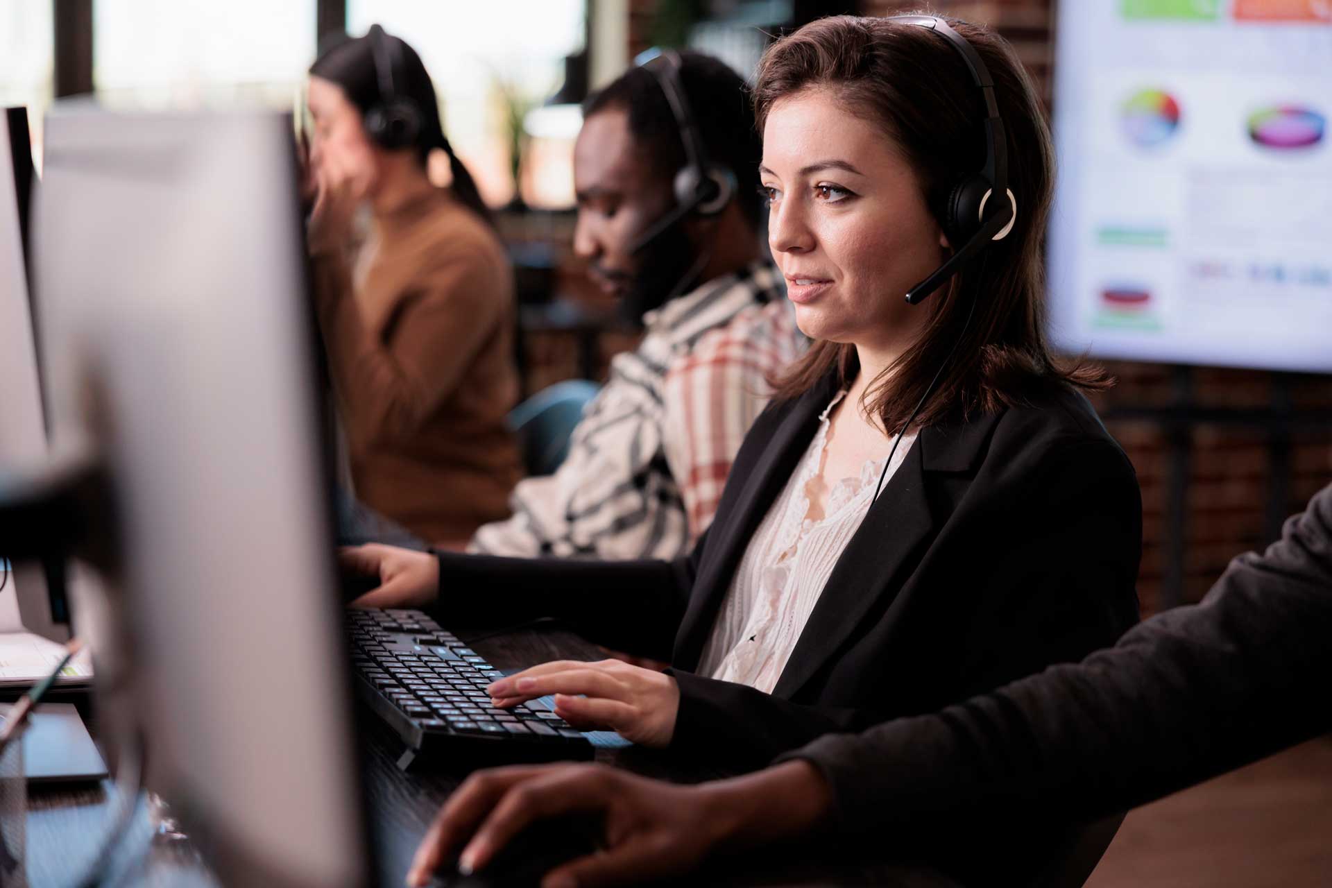 Three customer service representatives work at computers, wearing headsets in an office.