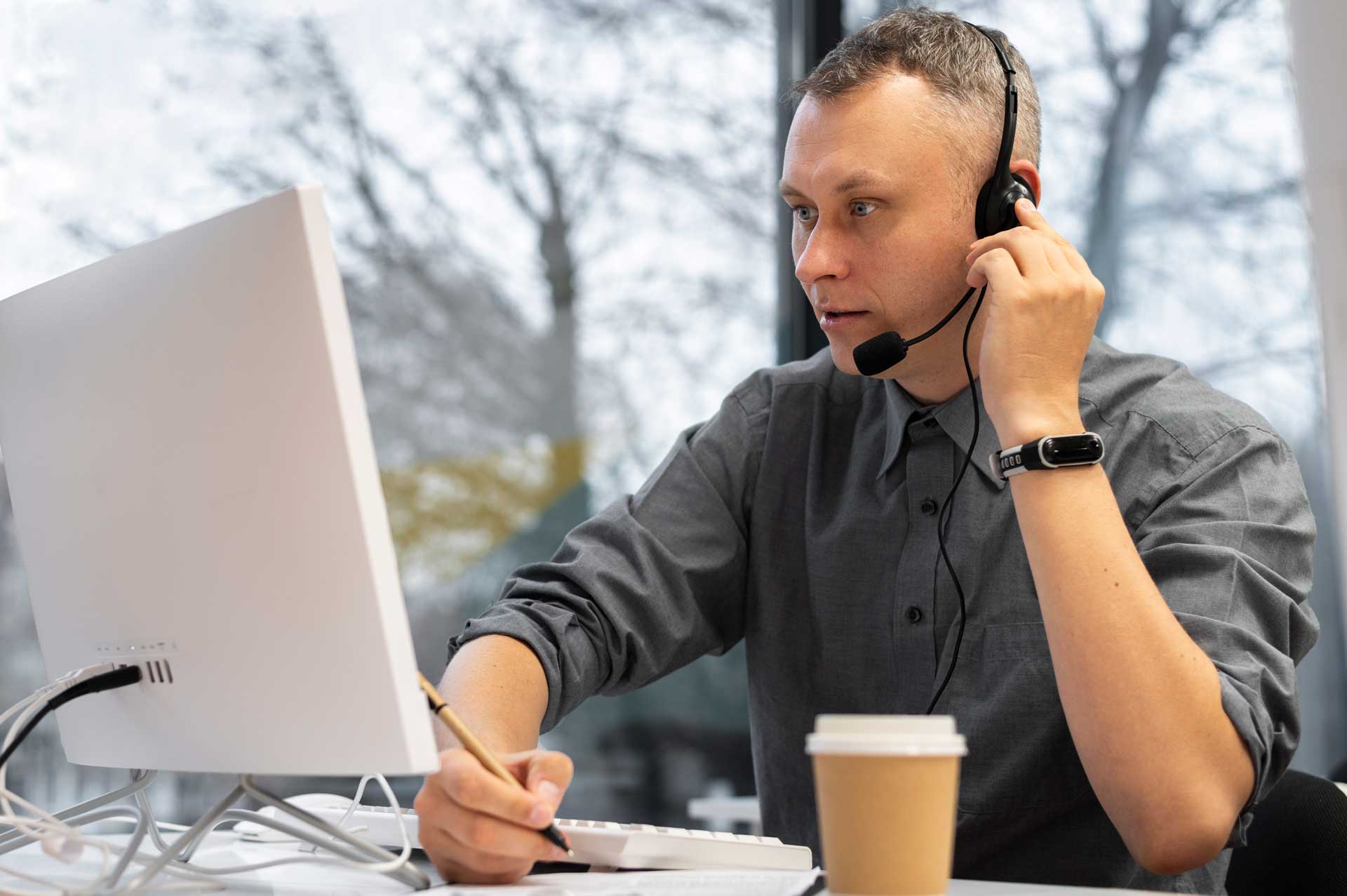 Man wearing a headset works on a computer, holding a pencil. A coffee cup is nearby.