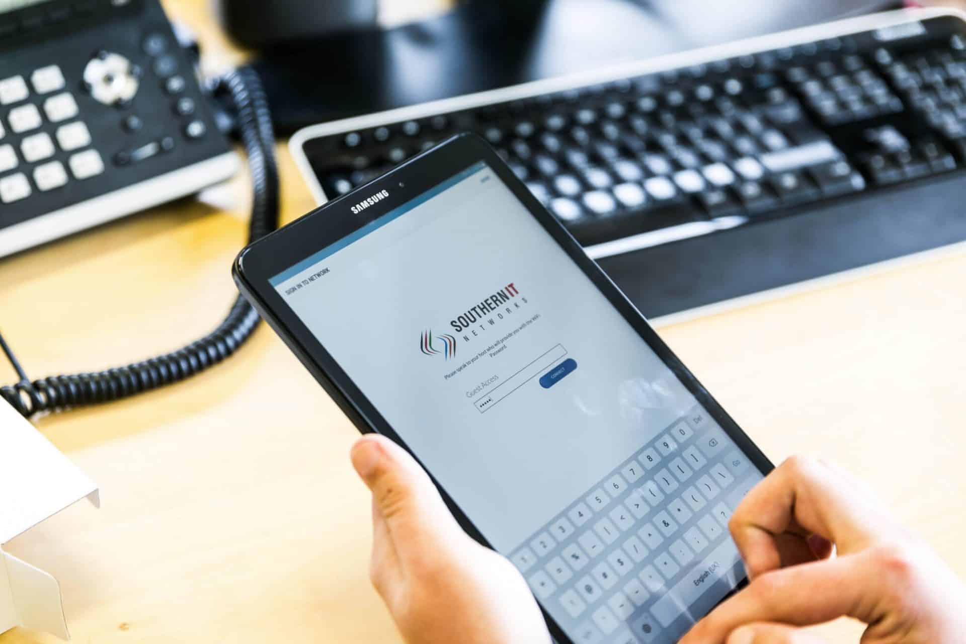 A person uses a Samsung tablet with a login screen, near a keyboard and phone.