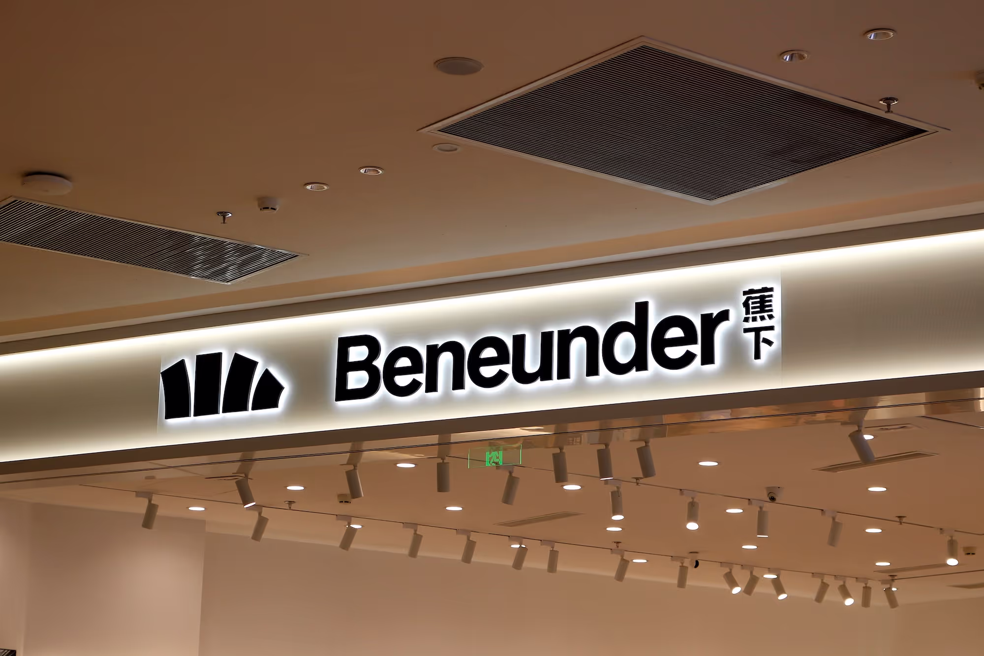 Illuminated store sign with the name 'Beneunder' and a black logo featuring four curved shapes.