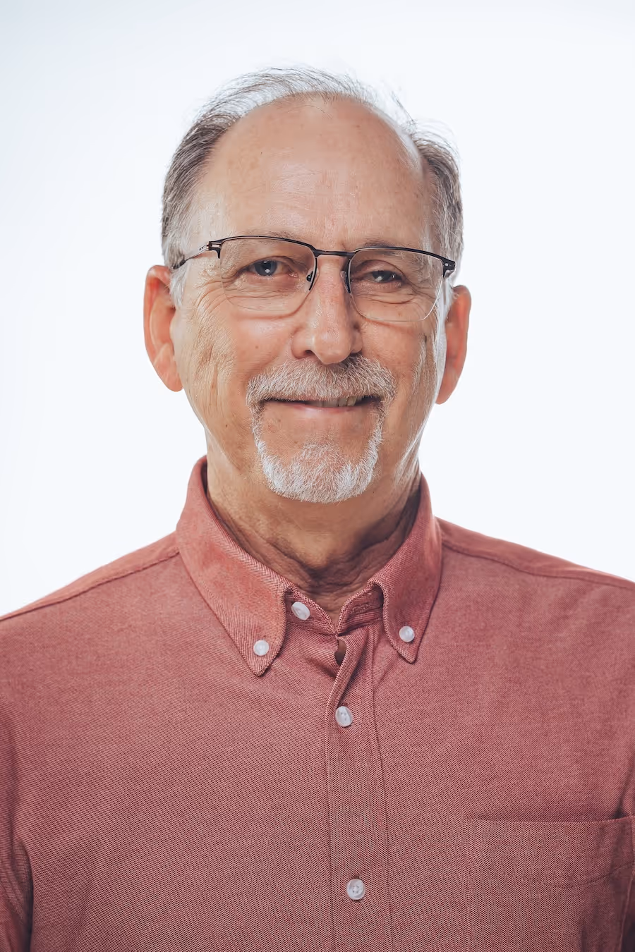 Smiling older man with glasses and gray goatee wearing a red button-up shirt against a white background.