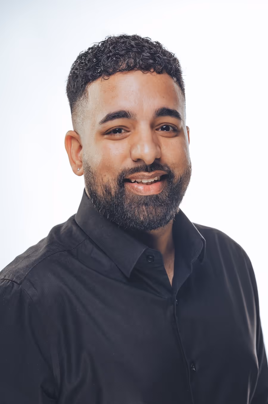 Smiling man with curly hair, beard, and black shirt against a light background.