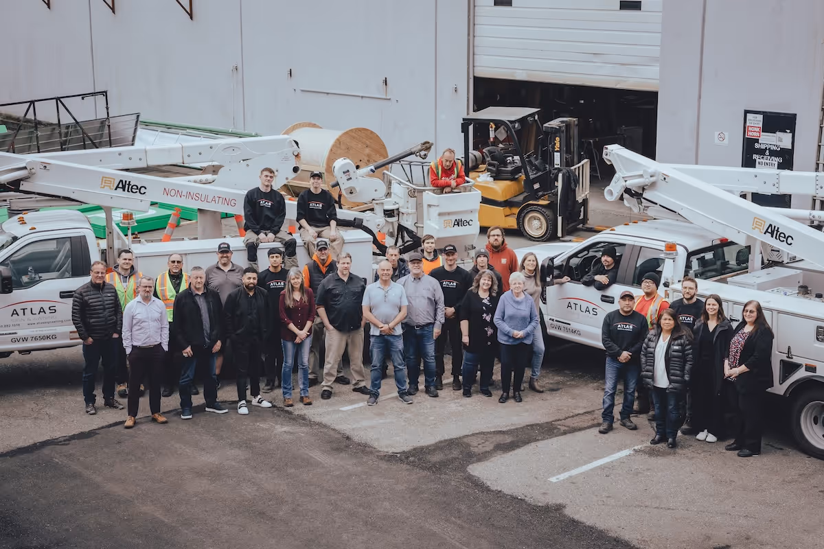 Group of about 30 people standing and sitting around two white utility trucks with Altec booms in an industrial parking lot.