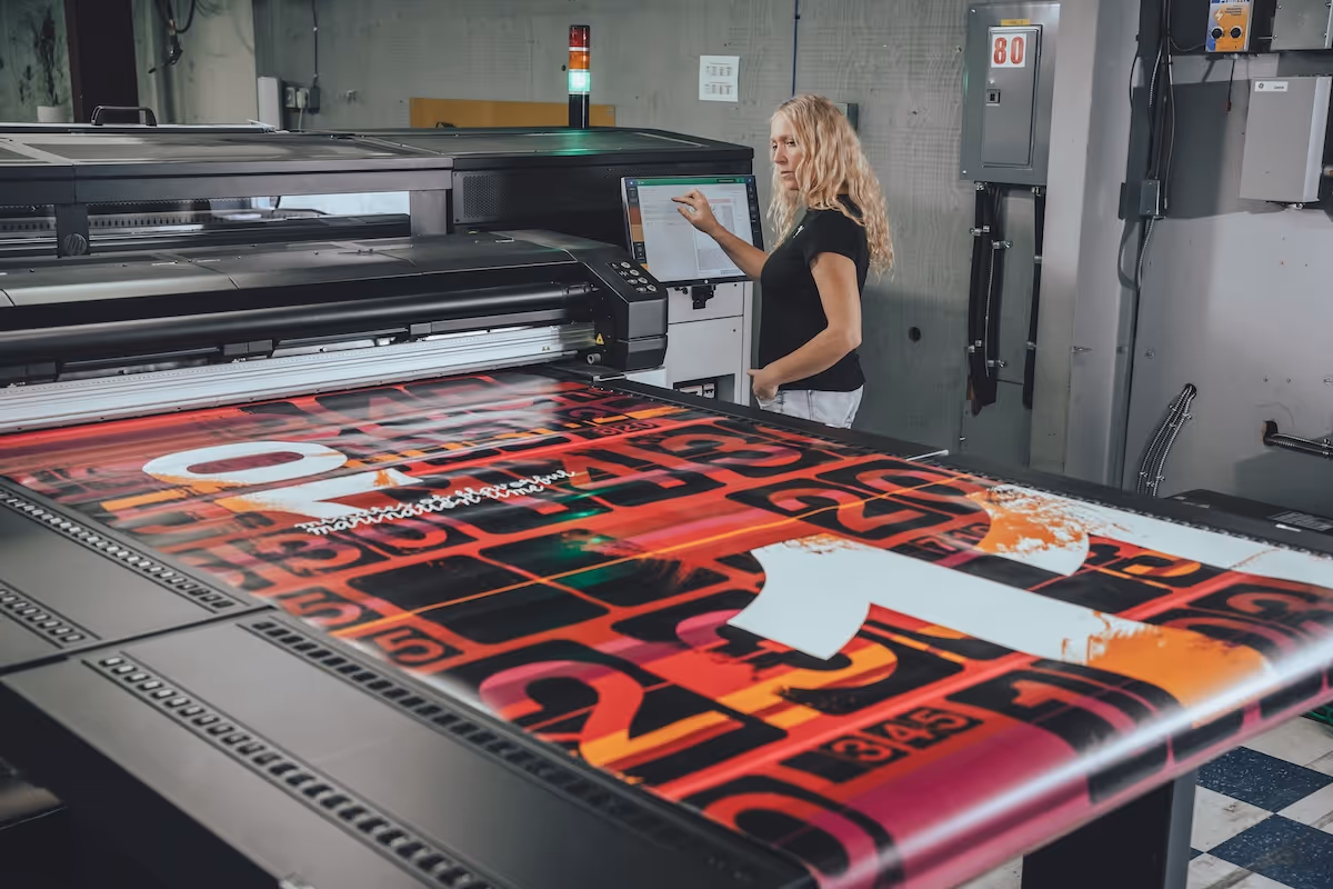 Woman operating a large-format printer producing a colorful graphic print with red, black, and orange tones.