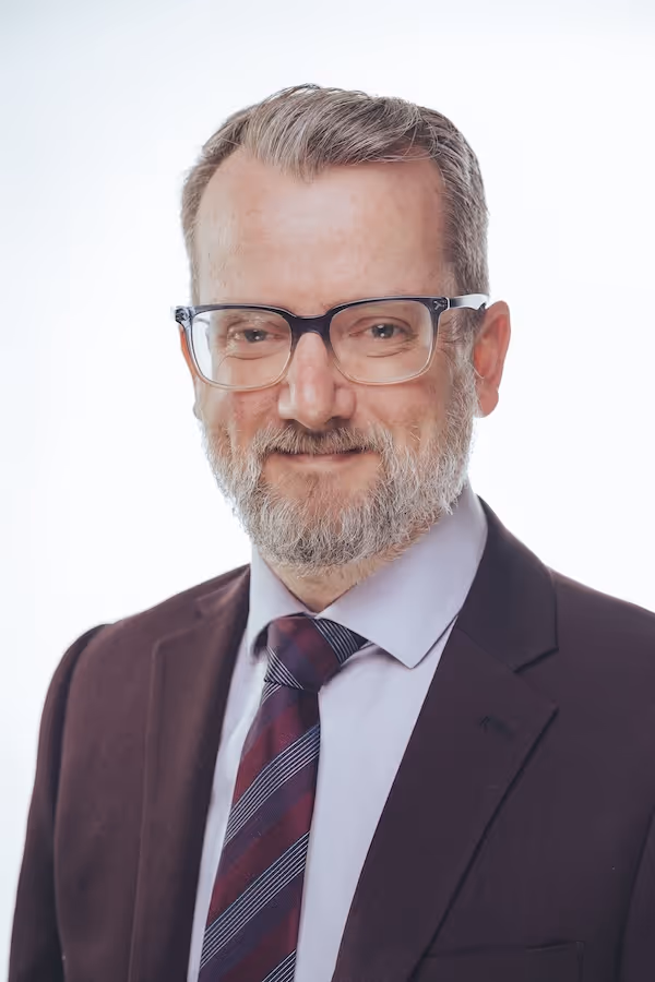 Smiling middle-aged man with gray hair, beard, glasses, wearing a dark suit and striped tie.