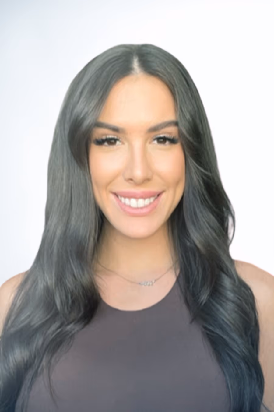 Smiling woman with long black wavy hair wearing a dark sleeveless top against a plain light background.