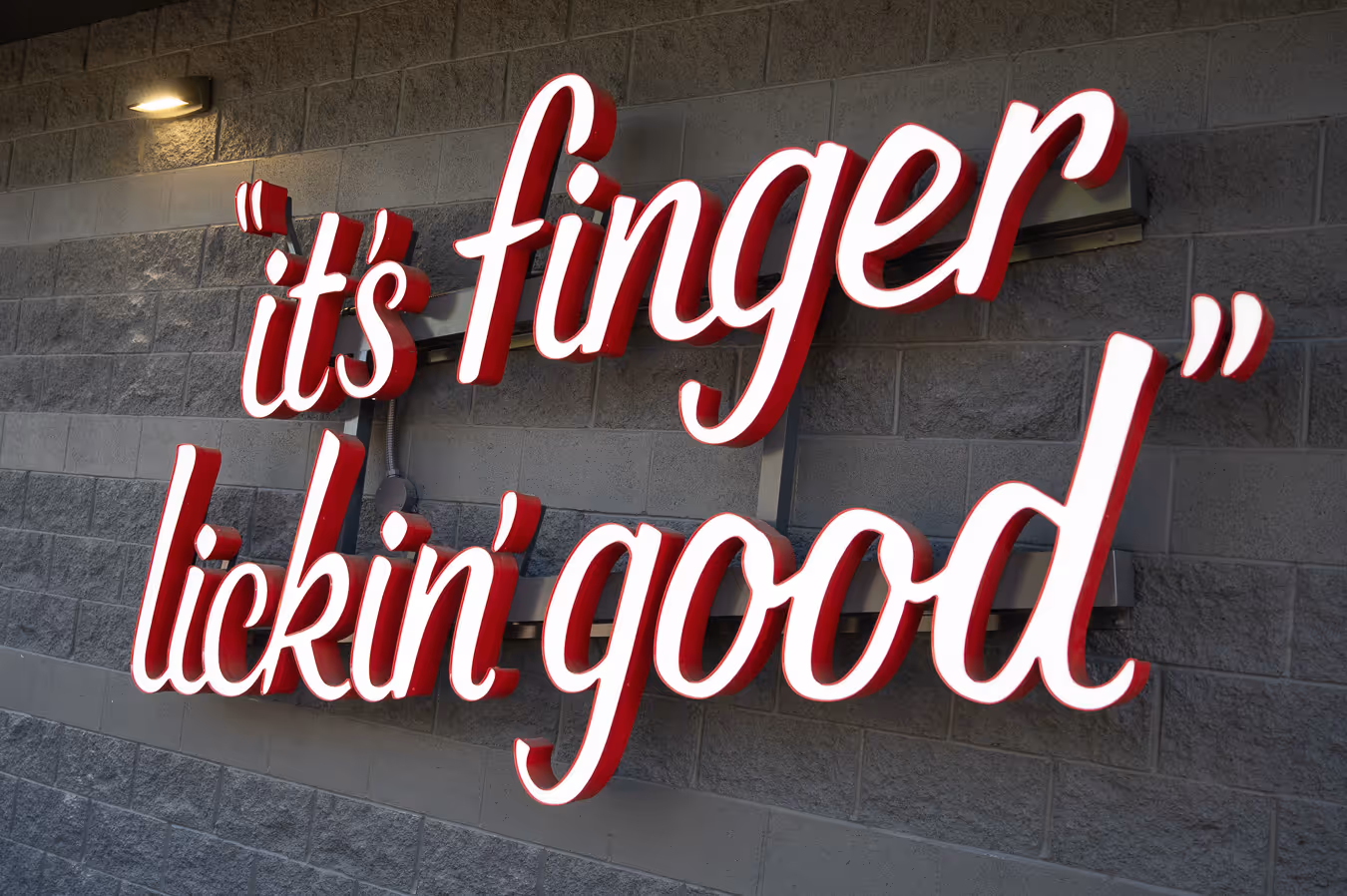 Illuminated sign on a gray brick wall reading 'it's finger lickin' good' in cursive script.