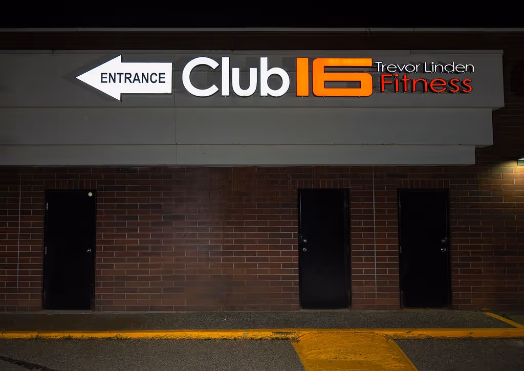 Illuminated sign for Club16 Trevor Linden Fitness with a left arrow labeled Entrance above three black doors on a brick wall at night.