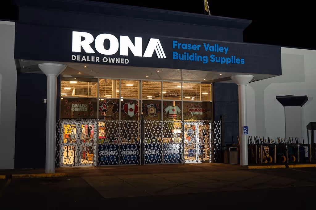 Night view of RONA Fraser Valley Building Supplies storefront with illuminated signage and closed security gates.