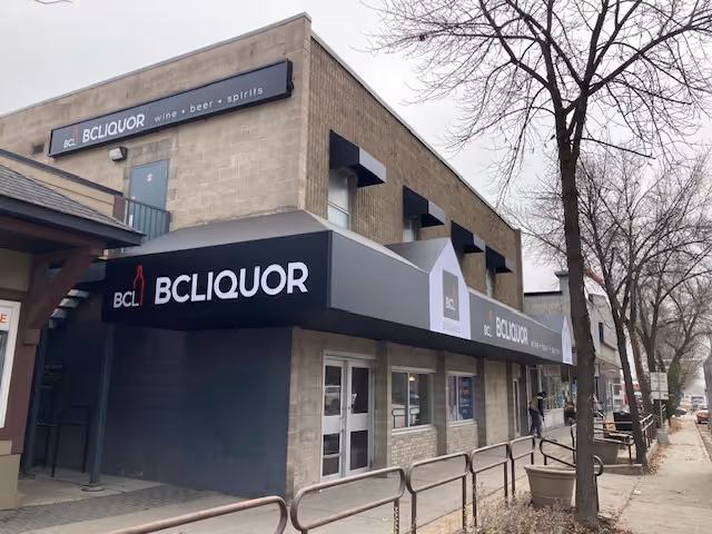 Street view of a corner building housing BCLiquor, a store specializing in wine, beer, and spirits, with a cloudy sky and leafless trees nearby.
