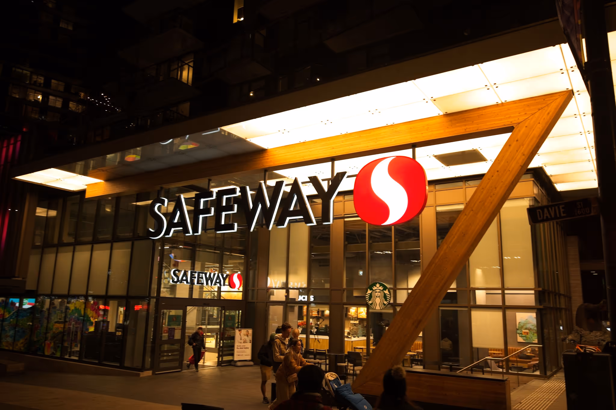Safeway grocery store entrance at night with illuminated signage and people walking nearby.