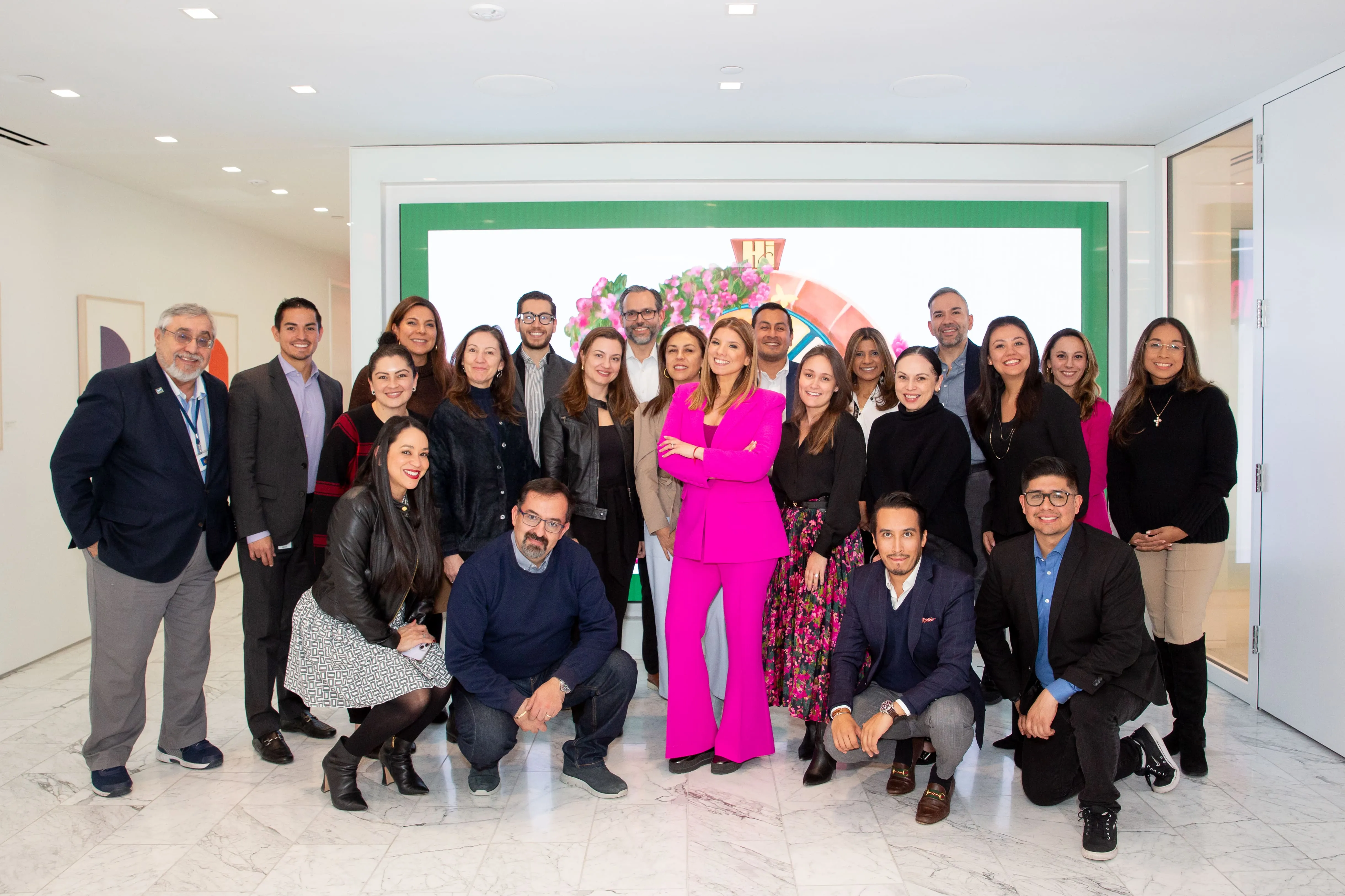 Group of 21 diverse people, mostly smiling, posing indoors on a marble floor with woman in bright pink suit centered.