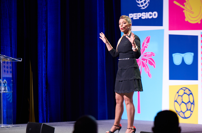 Woman in a black dress speaking on stage with PepsiCo logo and colorful graphics in the background.