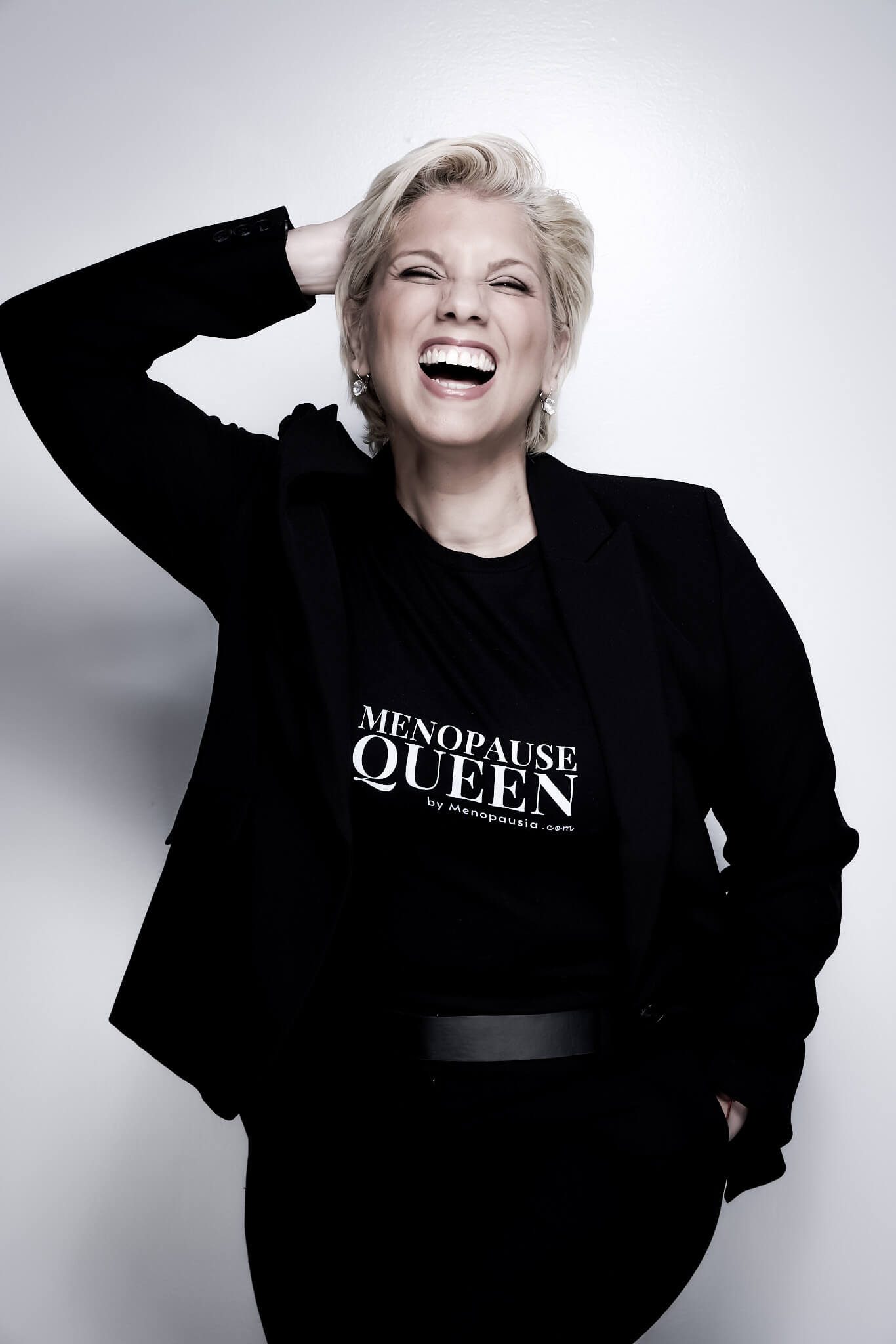 Woman with short blonde hair laughing, wearing a black blazer and a black shirt that reads 'MENOPAUSE QUEEN by Menopausia.com'.