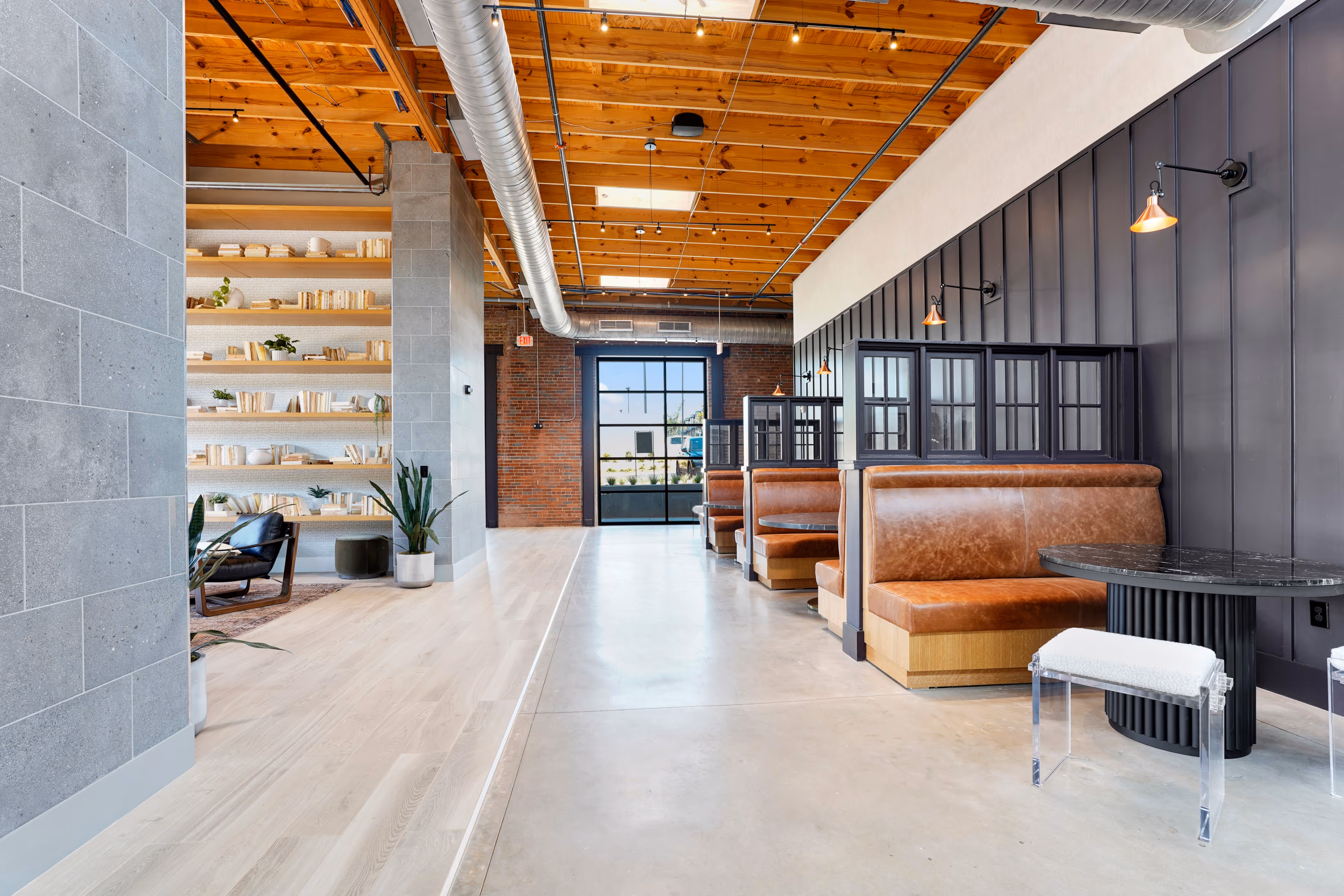 Modern interior with leather booth seating, black tables, wooden ceiling, and open shelves with books and plants.