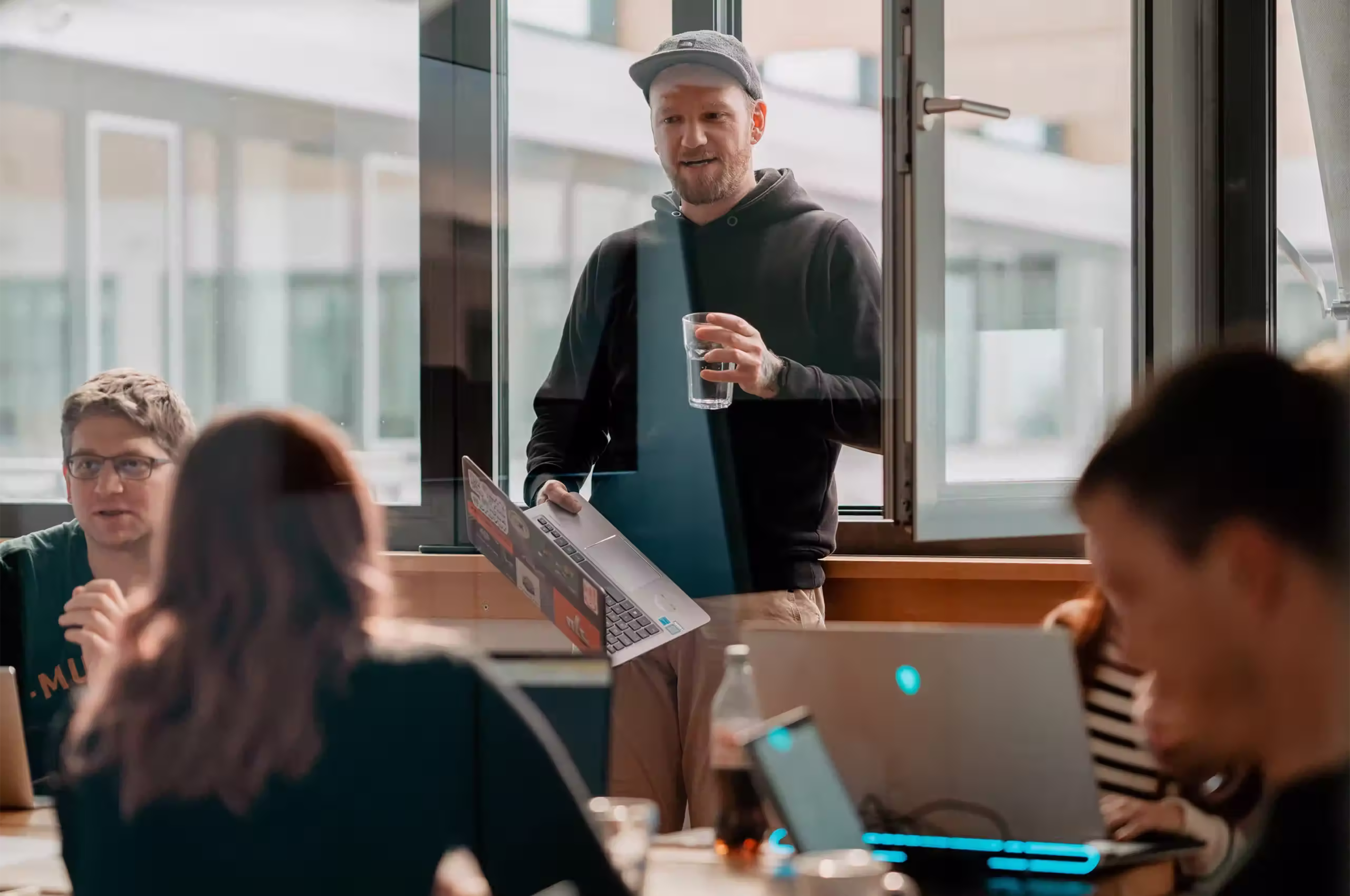 Ein Mann in schwarzer Kapuzenjacke und Kappe hält einen Laptop und ein Glas Wasser vor einem offenen Fenster, während andere Personen an Tischen mit Laptops sitzen.