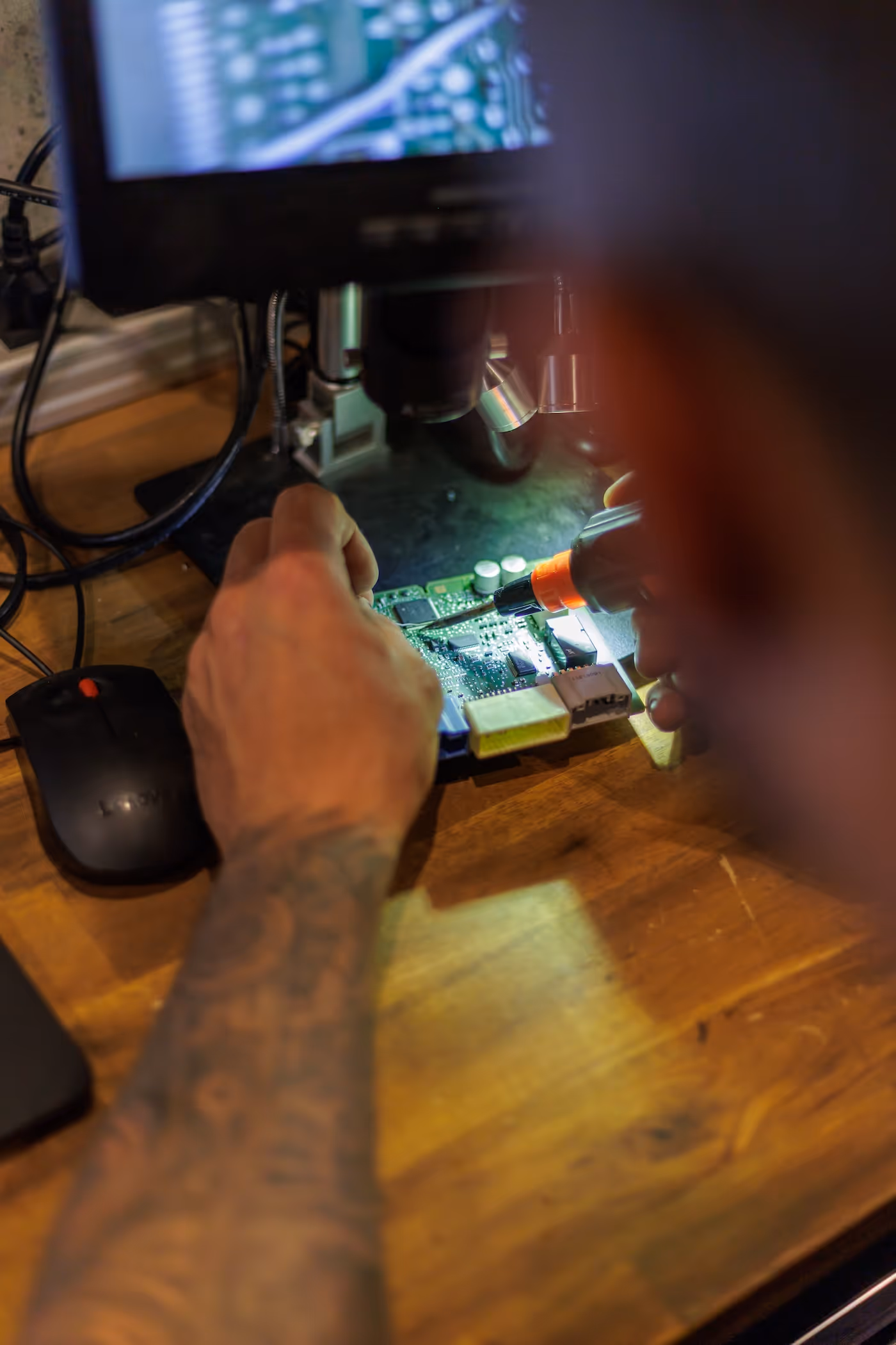 Person repairing a circuit board using a soldering tool under a microscope on a wooden desk.