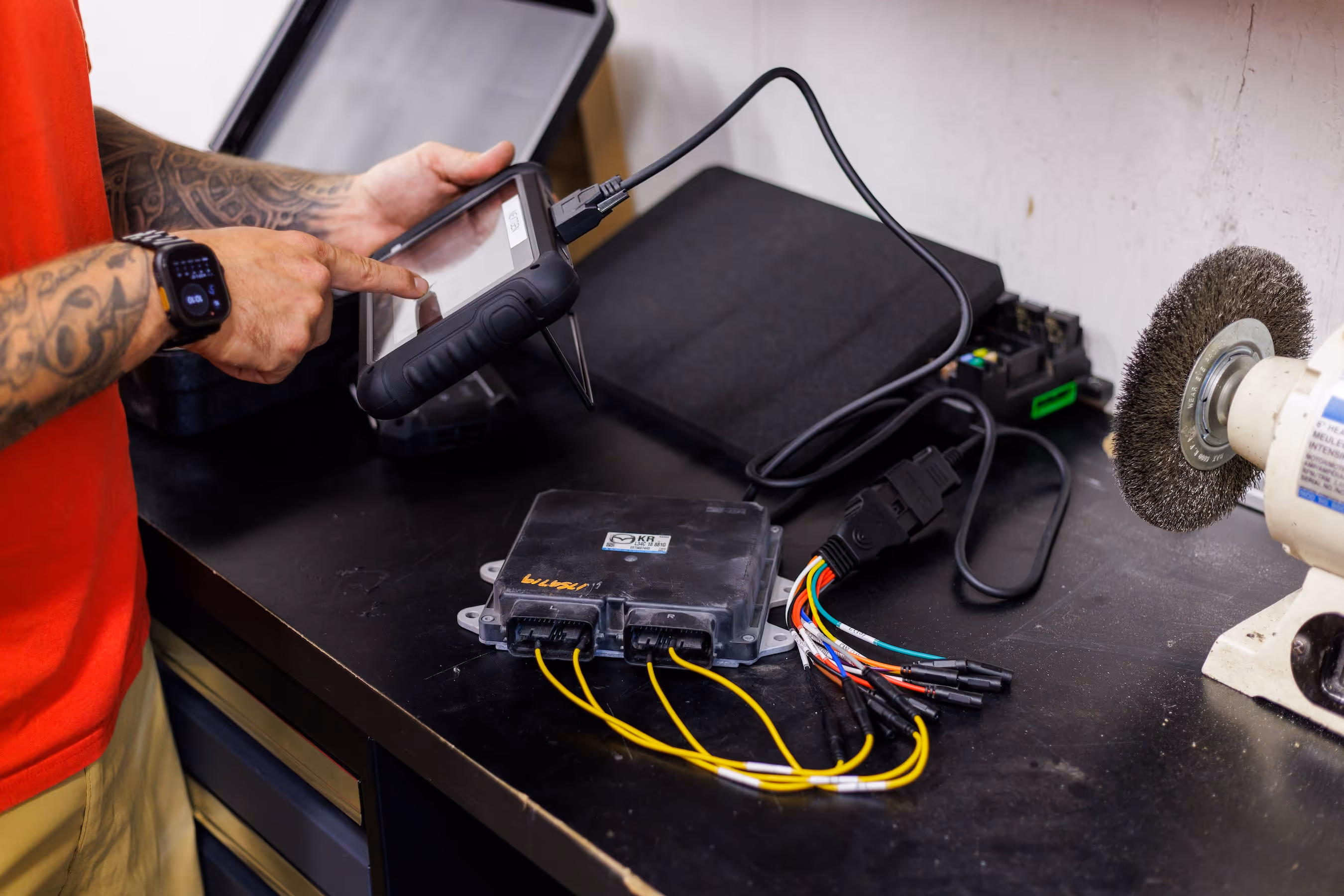Person with tattooed arm using a diagnostic tablet connected to a car ECU on a workbench.