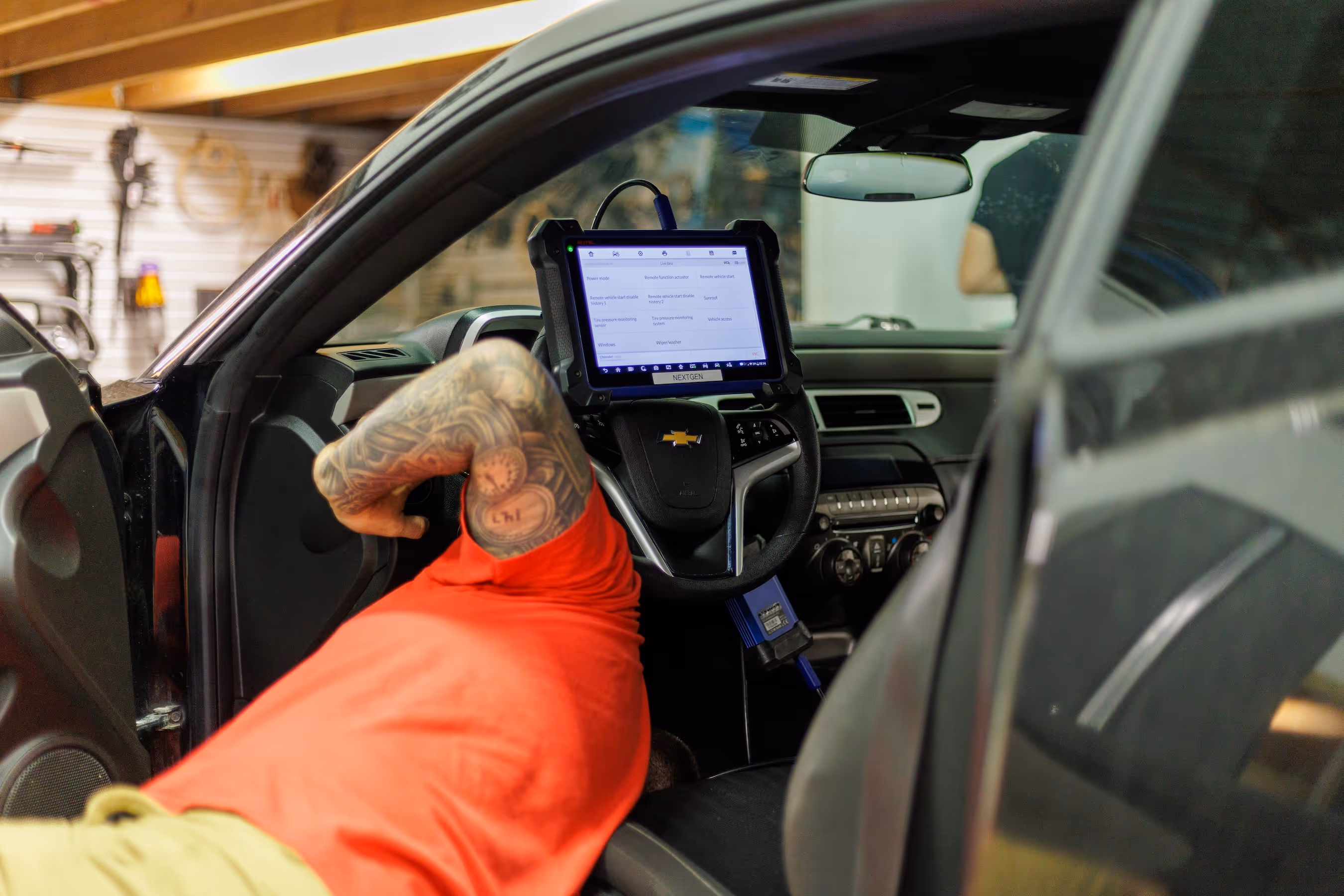 Person with tattooed arm in red shirt using a diagnostic tablet connected to a Chevrolet car inside a garage.
