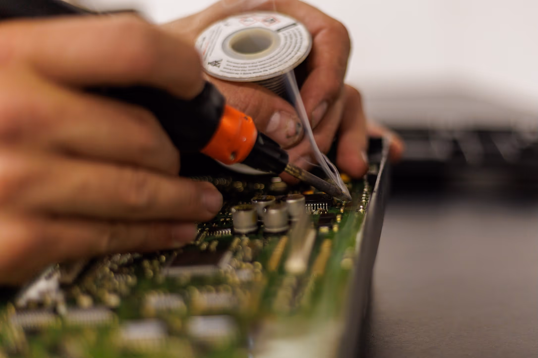 Hands soldering components on a green electronic circuit board using a soldering iron and solder wire.