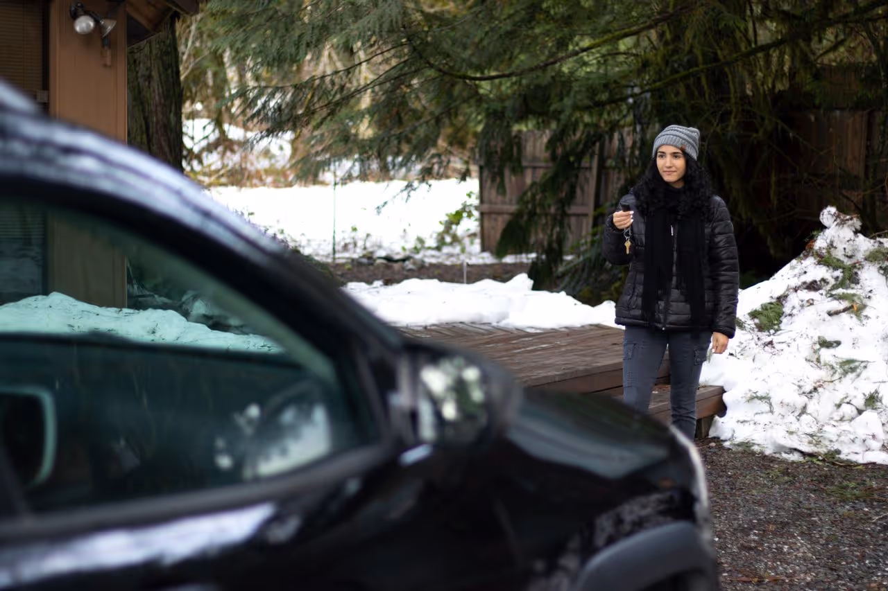Woman wearing winter clothes and a gray beanie holding car keys near a black car with snow and trees in the background.