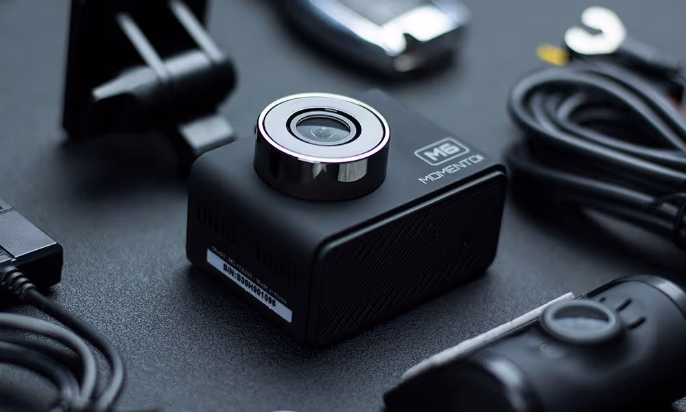 Black M6 Momentci action camera with lens on top surrounded by cables and accessories on a dark surface.