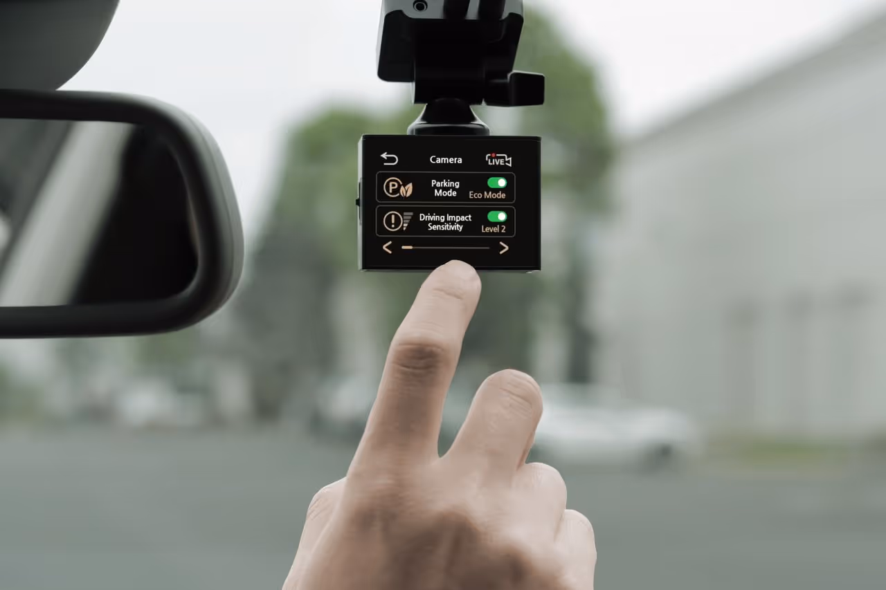 Hand adjusting settings on a car dash camera displaying Parking Mode in Eco Mode and Driving Impact Sensitivity at Level 2.