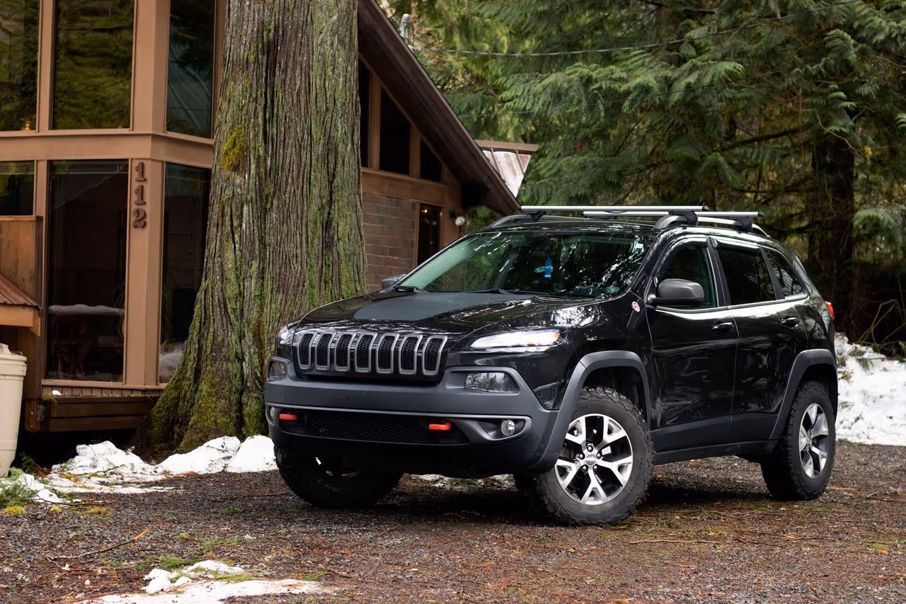 Black Jeep Cherokee SUV parked on a gravel driveway in front of a cabin with large tree trunk and green forest background.