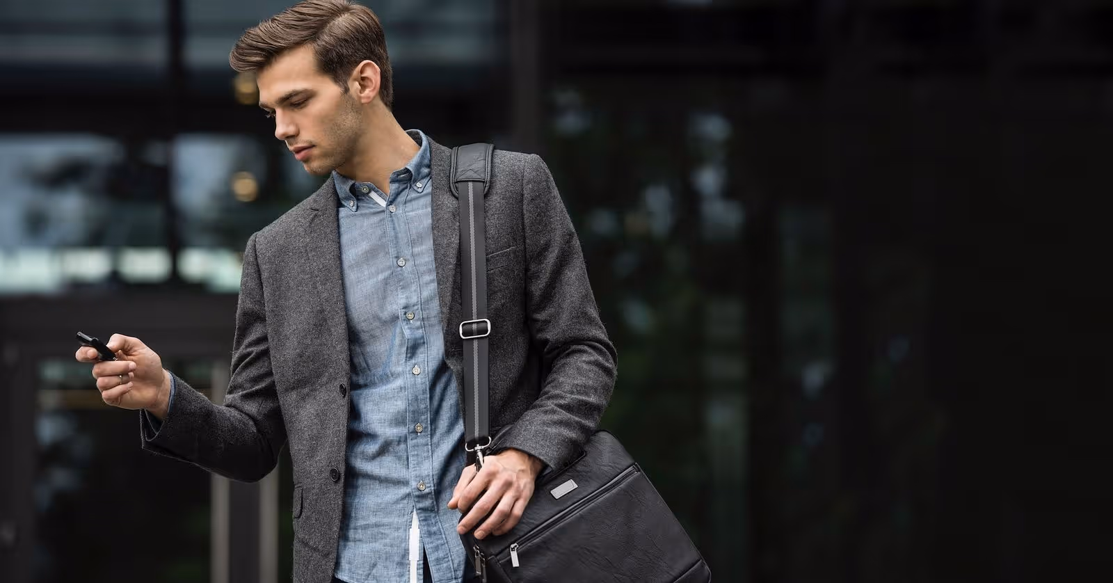 Young man in a grey blazer and blue shirt holding a smartphone and carrying a black leather shoulder bag.