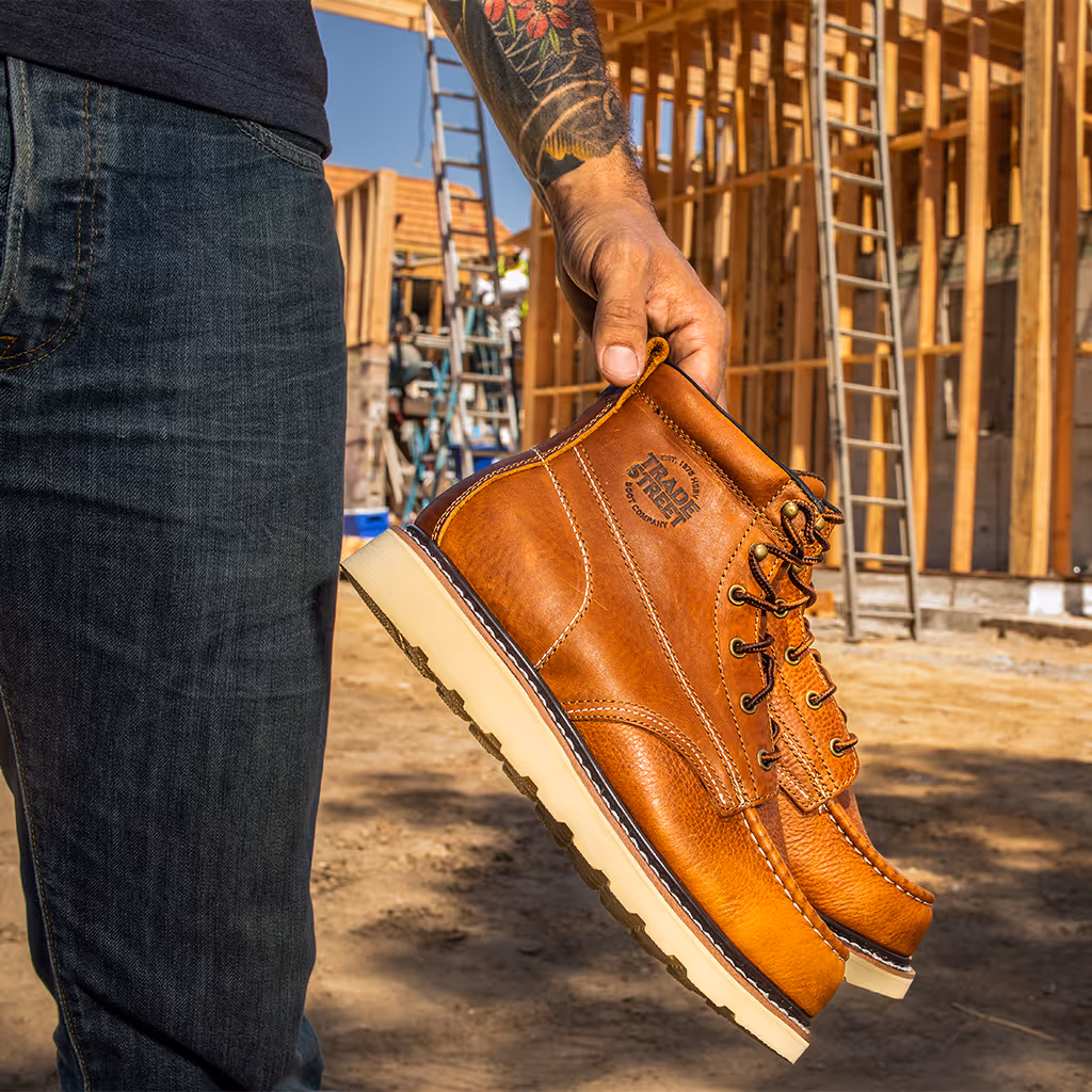 A worker holding a pair of Trade Street work boots in front of a construction site.