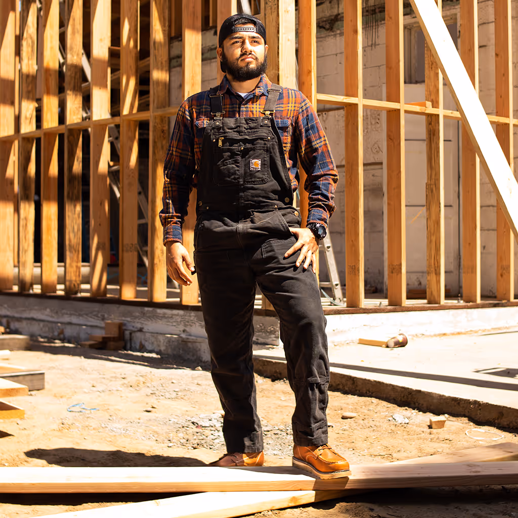 Construction worker standing confidently on a job site wearing black work overalls over a plaid shirt, a backward cap, and brown leather work boots, with exposed wooden framing in the background that conveys craftsmanship, durability, and hands-on workwear performance.