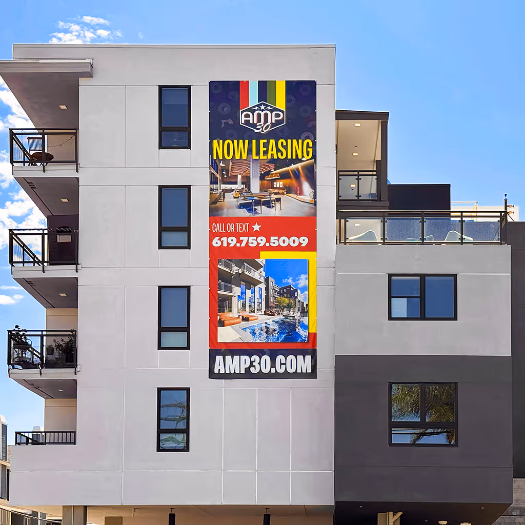 Exterior view of a modern apartment building with a large vertical AMP 30 banner mounted on the facade, featuring the AMP 30 logo, bold “Now Leasing” headline, interior and pool imagery, a call-or-text phone number (619-759-5009), and the website AMP30.com, set against a clear blue sky.