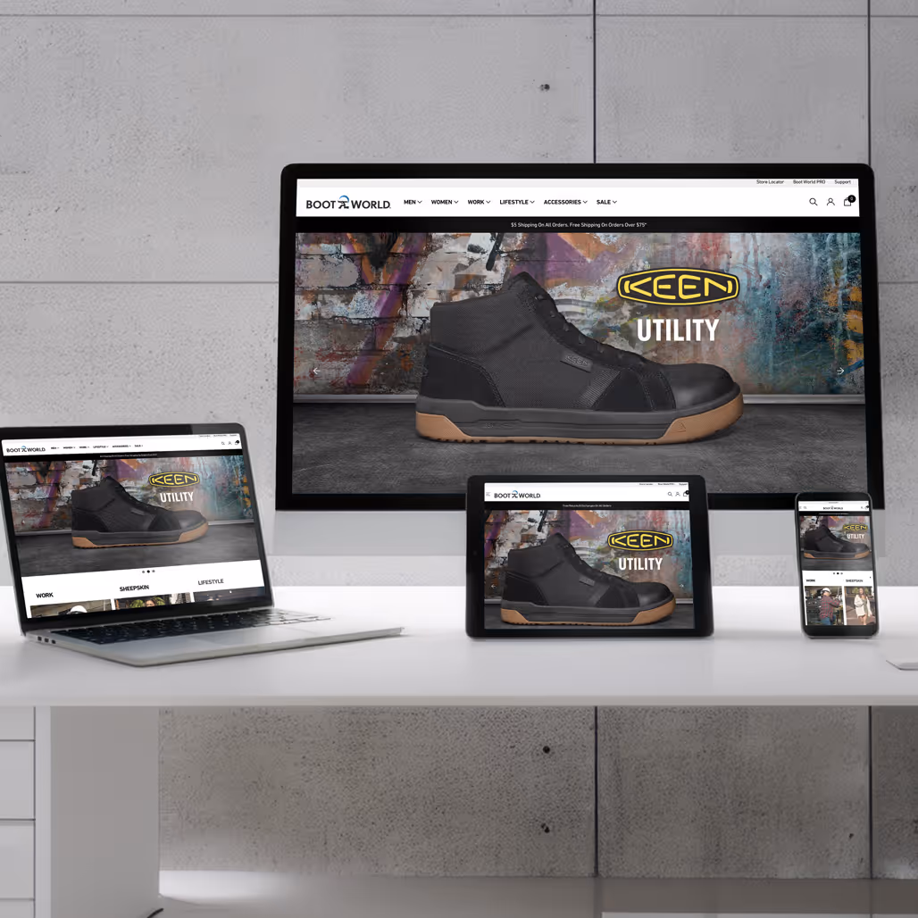 Boot World website displayed responsively across a desktop monitor, laptop, tablet, and smartphone on a white desk, featuring a Keen Utility work boot hero image against an urban graffiti-style background.