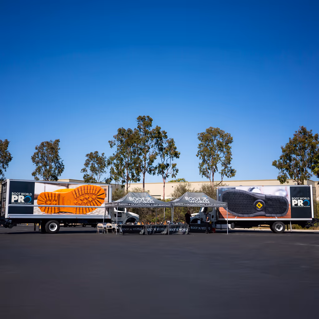 Outdoor Boot World PRO event setup featuring two branded box trucks with oversized boot sole graphics, a central canopy tent with Boot World signage, and a promotional booth arranged in a parking lot under a clear blue sky with trees and buildings in the background.