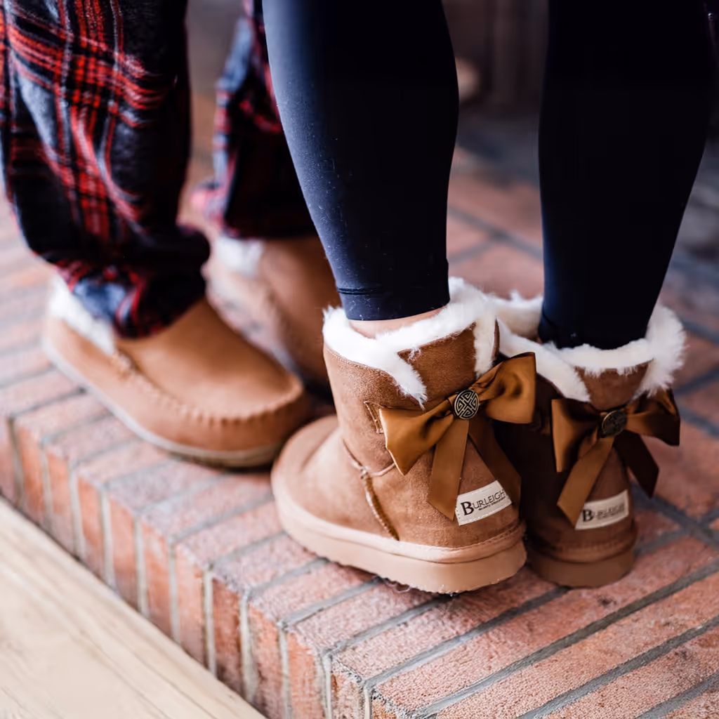 Two people standing on brick steps wearing cozy Burleigh sheepskin boots with soft fur lining and decorative bows, paired with leggings and plaid pants, creating a warm, casual lifestyle scene.