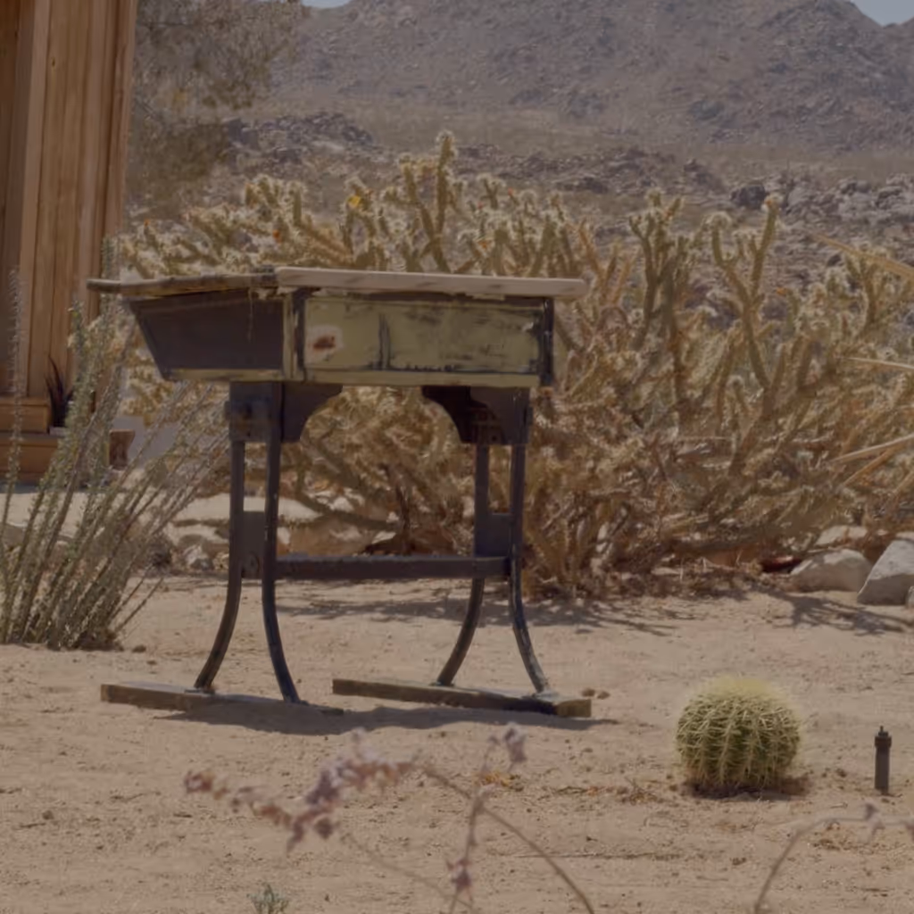 Weathered desk standing on curved legs in a desert landscape, surrounded by sandy ground, cacti, and scrub plants, with rocky hills and a rustic structure in the background under bright daylight.
