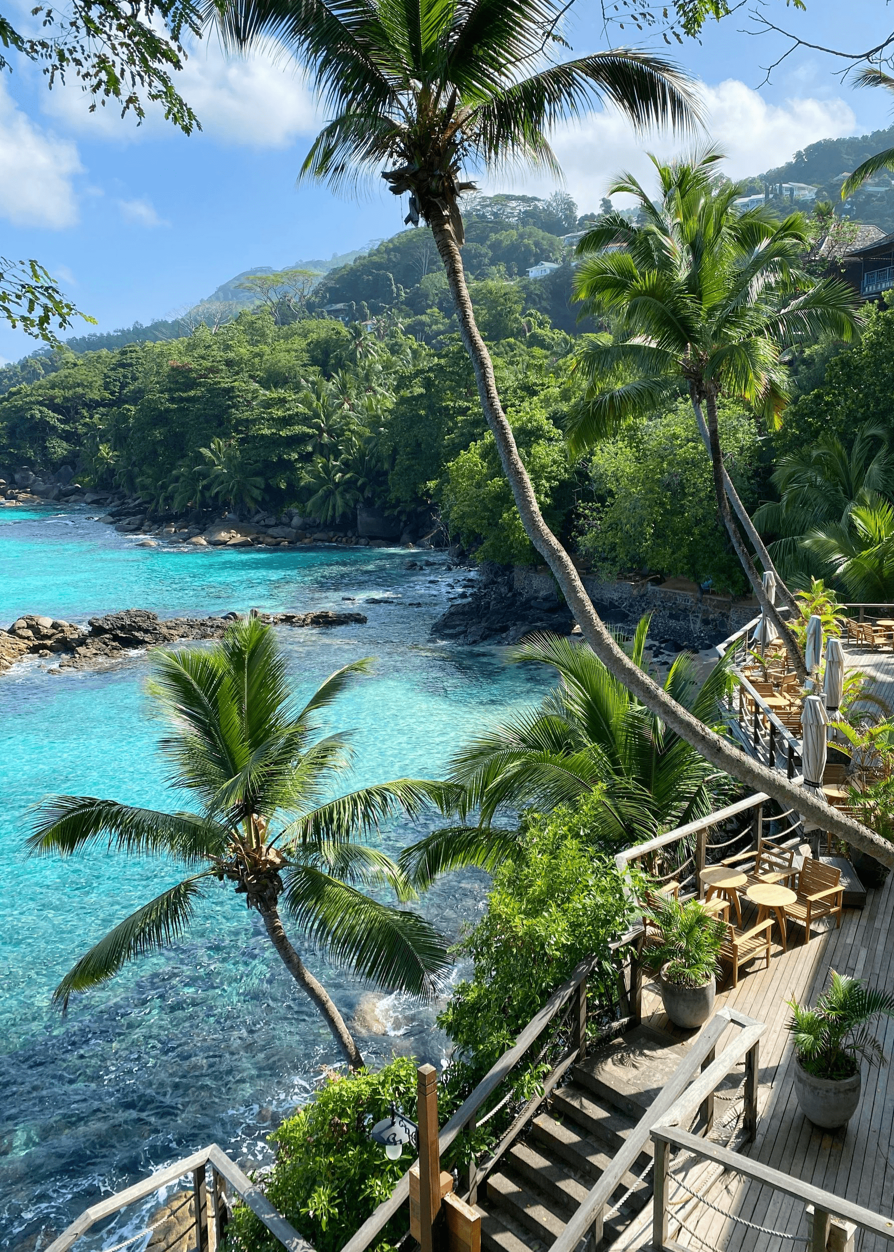 Blick auf eine tropische Bucht mit türkisblauem Wasser, Palmen und einer Holzterrasse mit Sitzgelegenheiten, eingebettet in üppige grüne Vegetation.