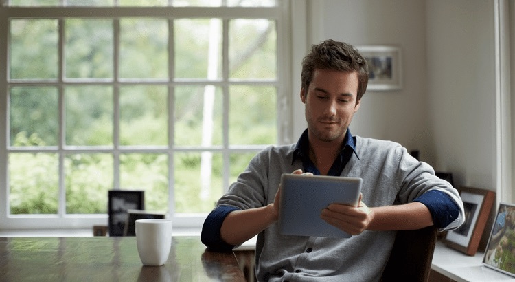 Man using a tablet at a kitchen table with a coffee mug in front of a bright window