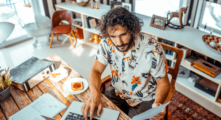Man reviewing paperwork while using a laptop at a wooden dining table.